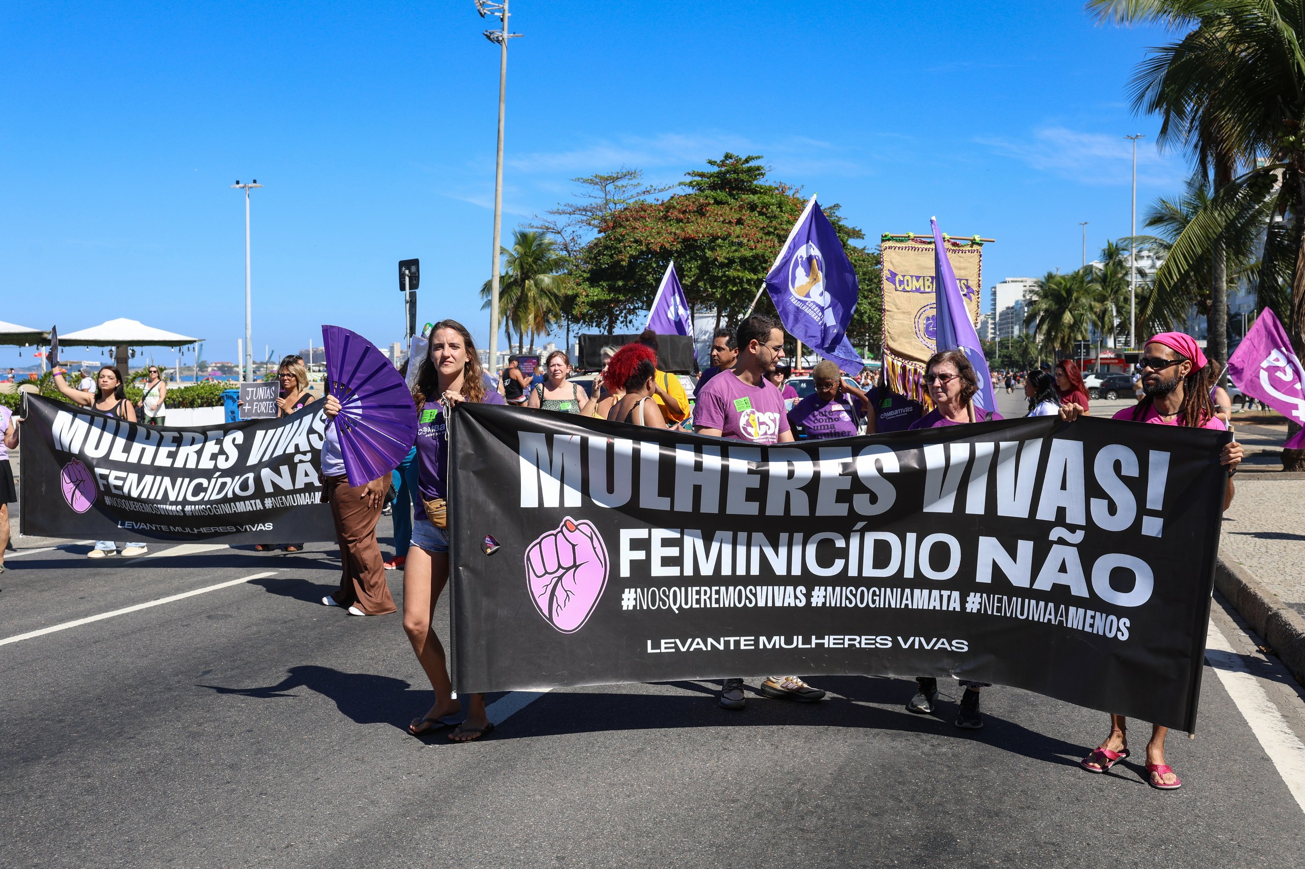 Grupo de mulheres segura faixa com a frase 'Mulheres Vivas! Feminicídio Não' durante manifestação em via pública.
