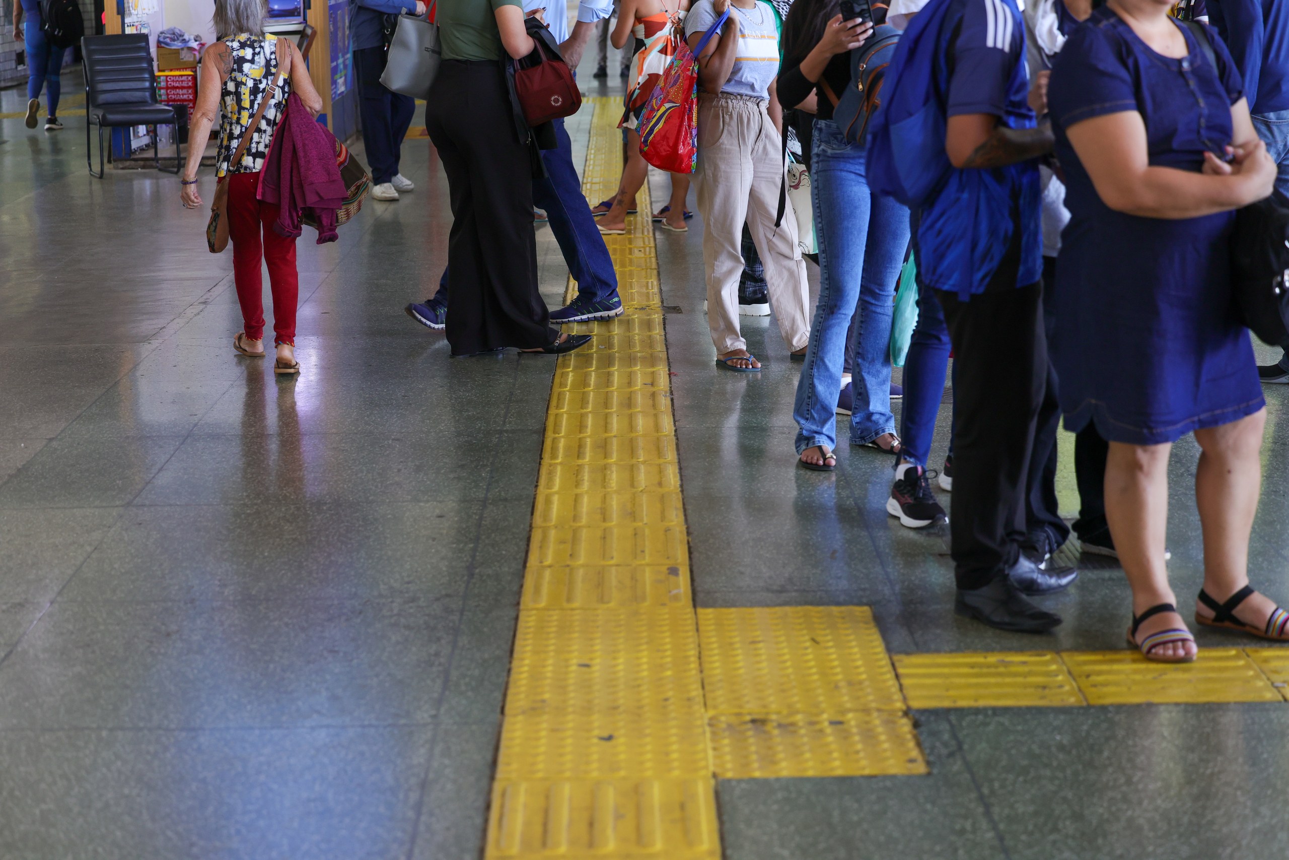 Pessoas aguardam em uma plataforma de transporte público com piso tátil amarelo.