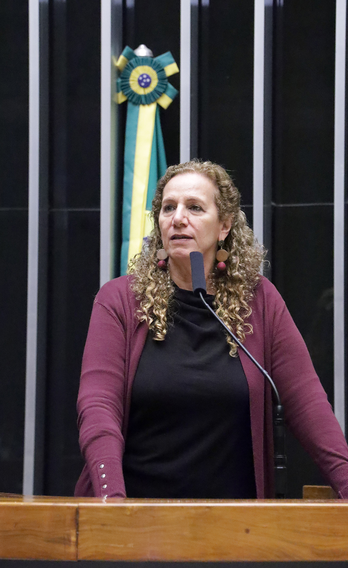 Mulher discursando em tribuna com microfone em ambiente formal.