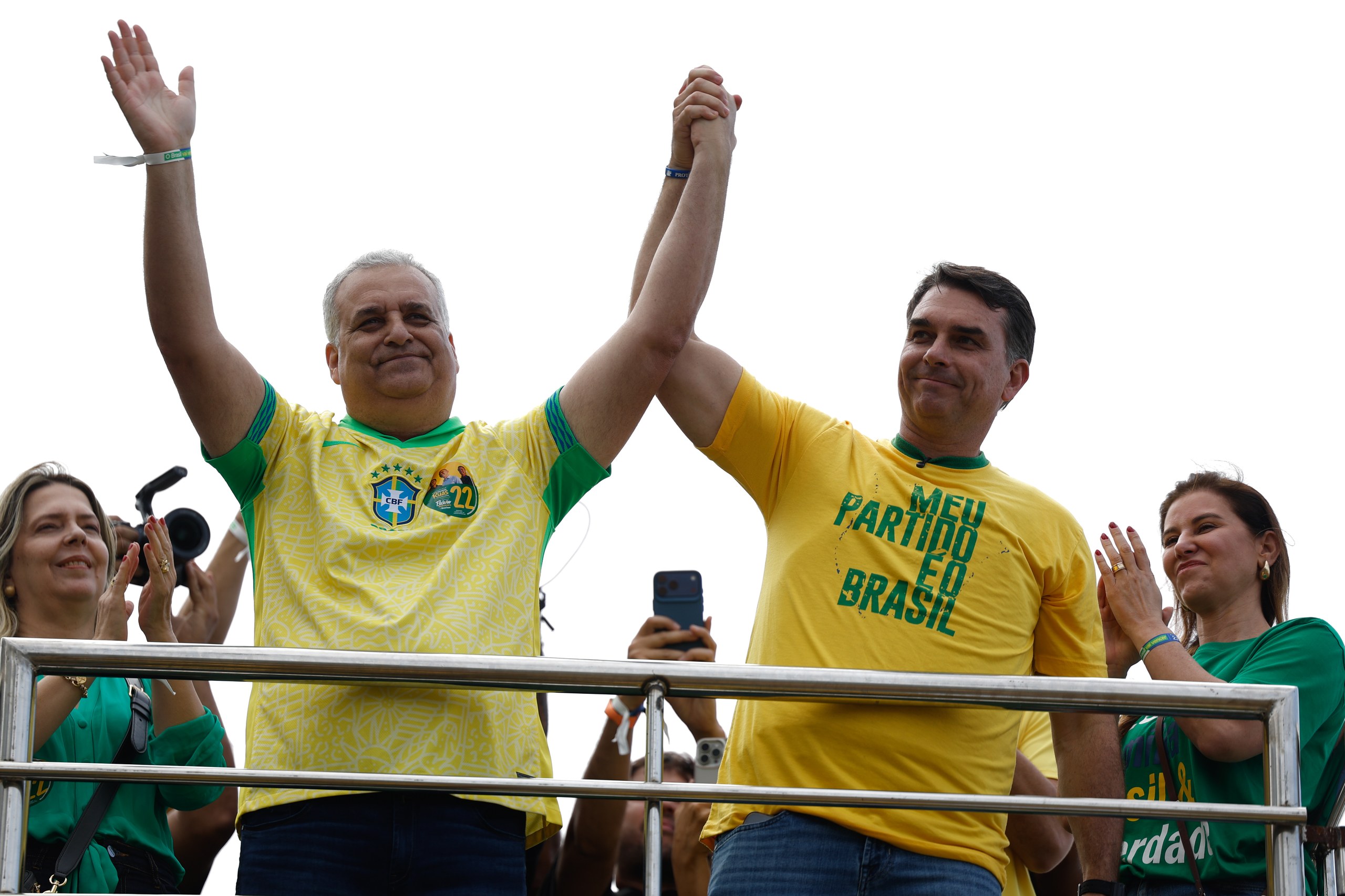 Dois homens com camisas amarelas celebram com as mãos erguidas durante um evento político ao ar livre.