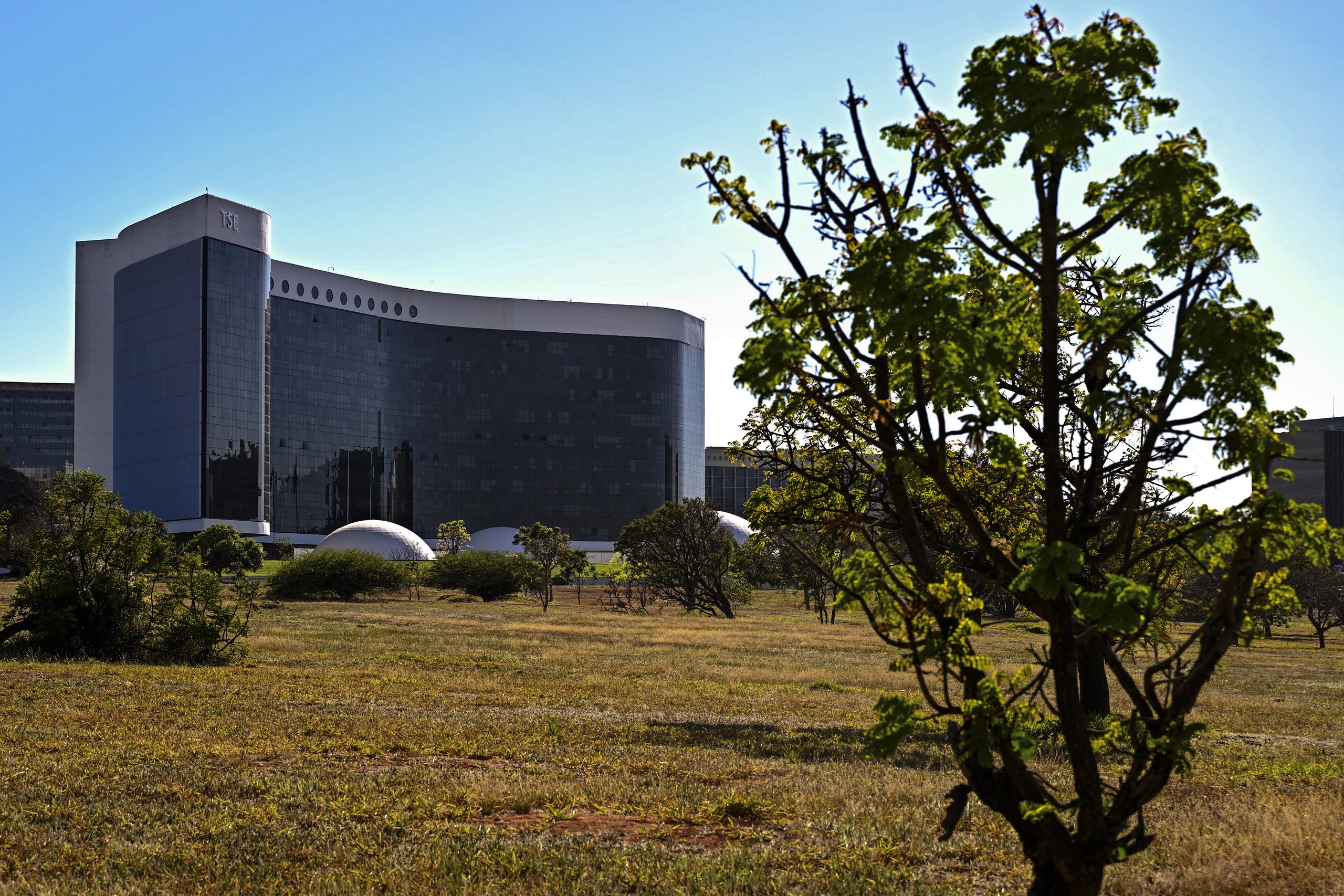 Vista externa de um edifício moderno de vidro cercado por áreas verdes sob um céu limpo.
