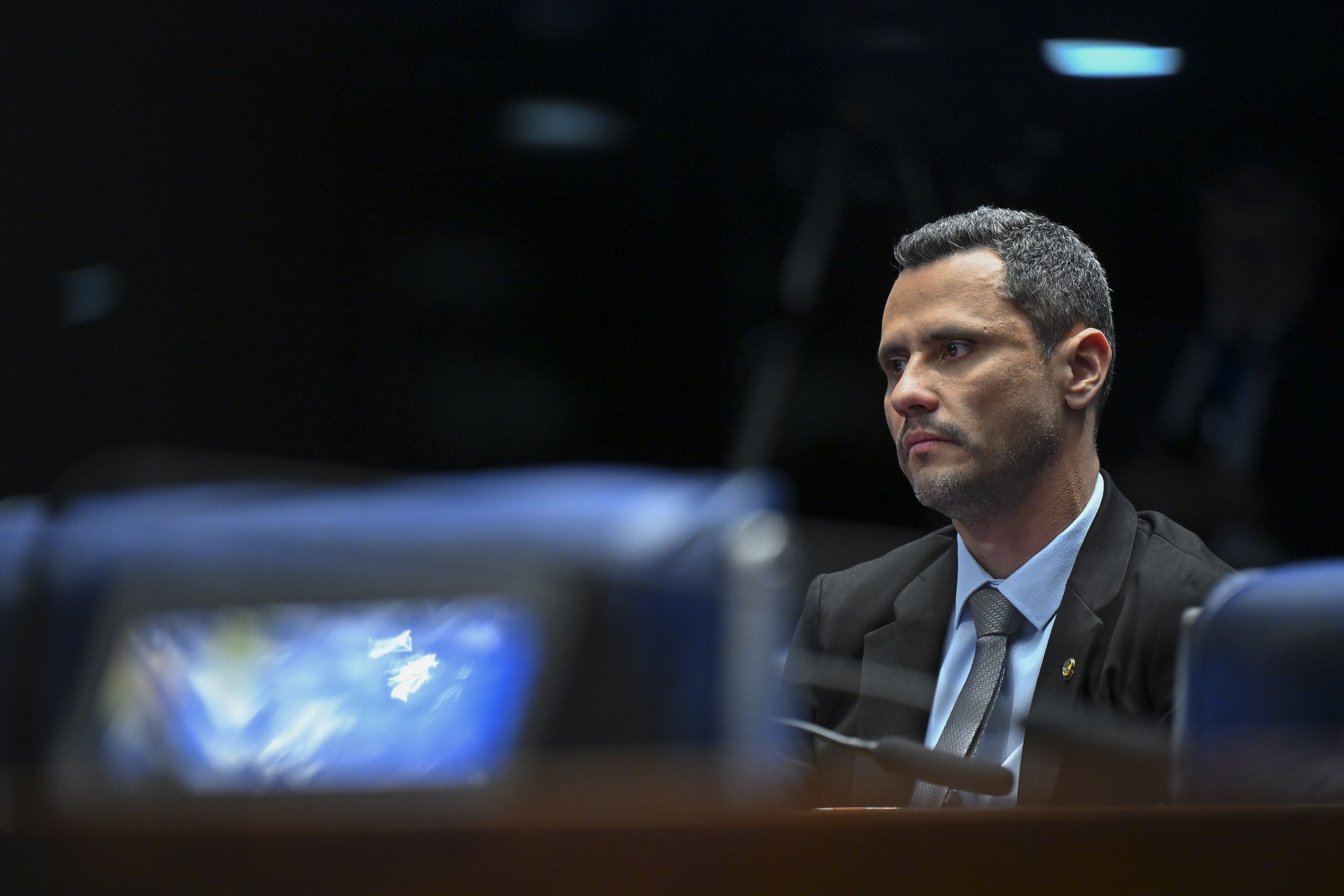 Homem vestindo terno e gravata com expressão séria em ambiente formal.