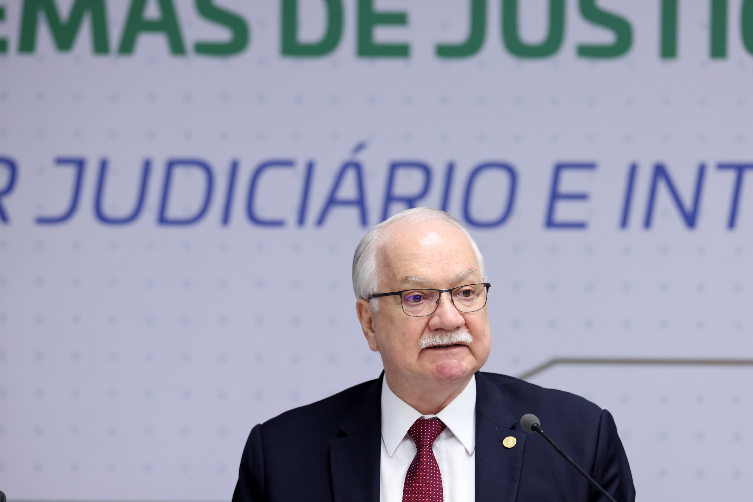 Um homem de terno e óculos fala ao microfone em frente a um painel com textos sobre o judiciário em um ambiente formal. Na foto: Edson Fachin.