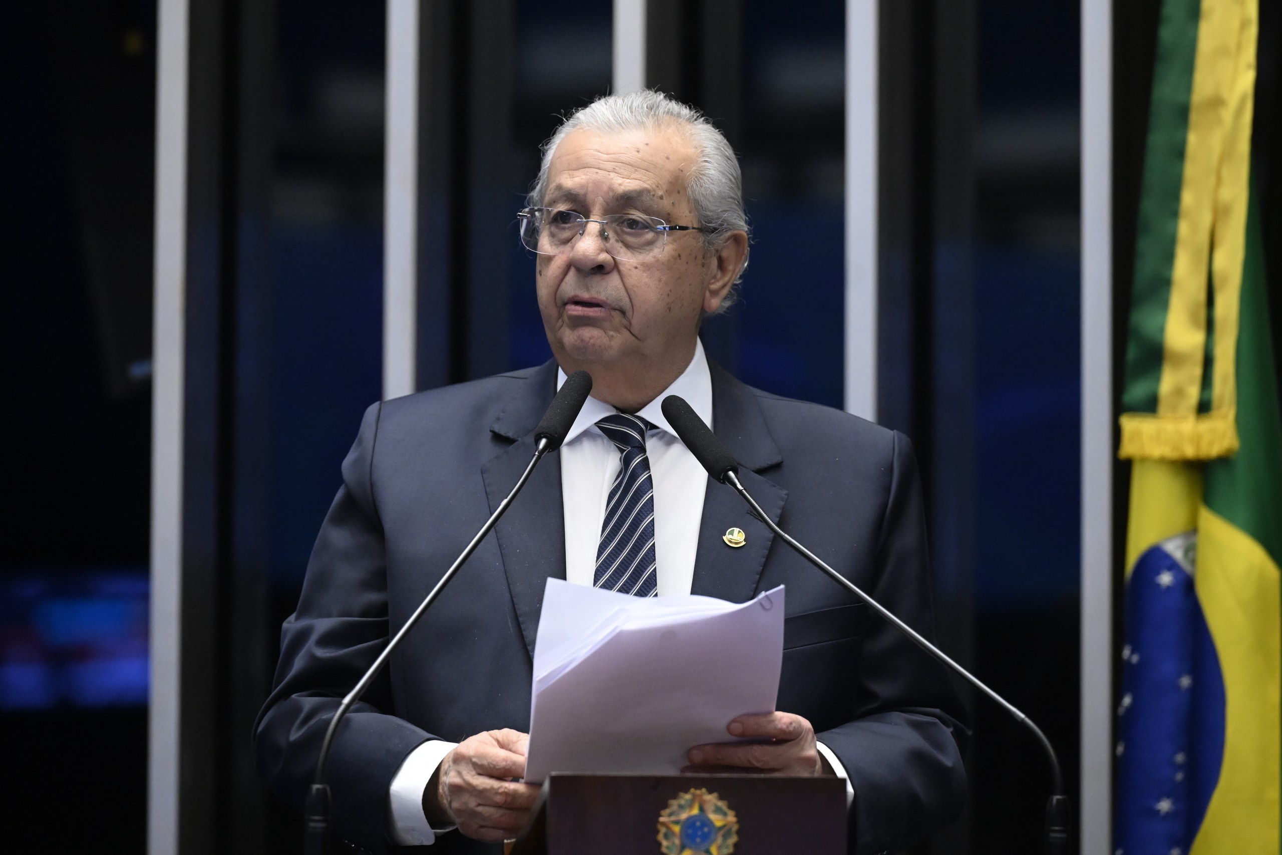 Um homem de terno e óculos lê um documento em um púlpito com microfones, em ambiente institucional com a bandeira do Brasil ao fundo. Na foto: Jayme Campos.