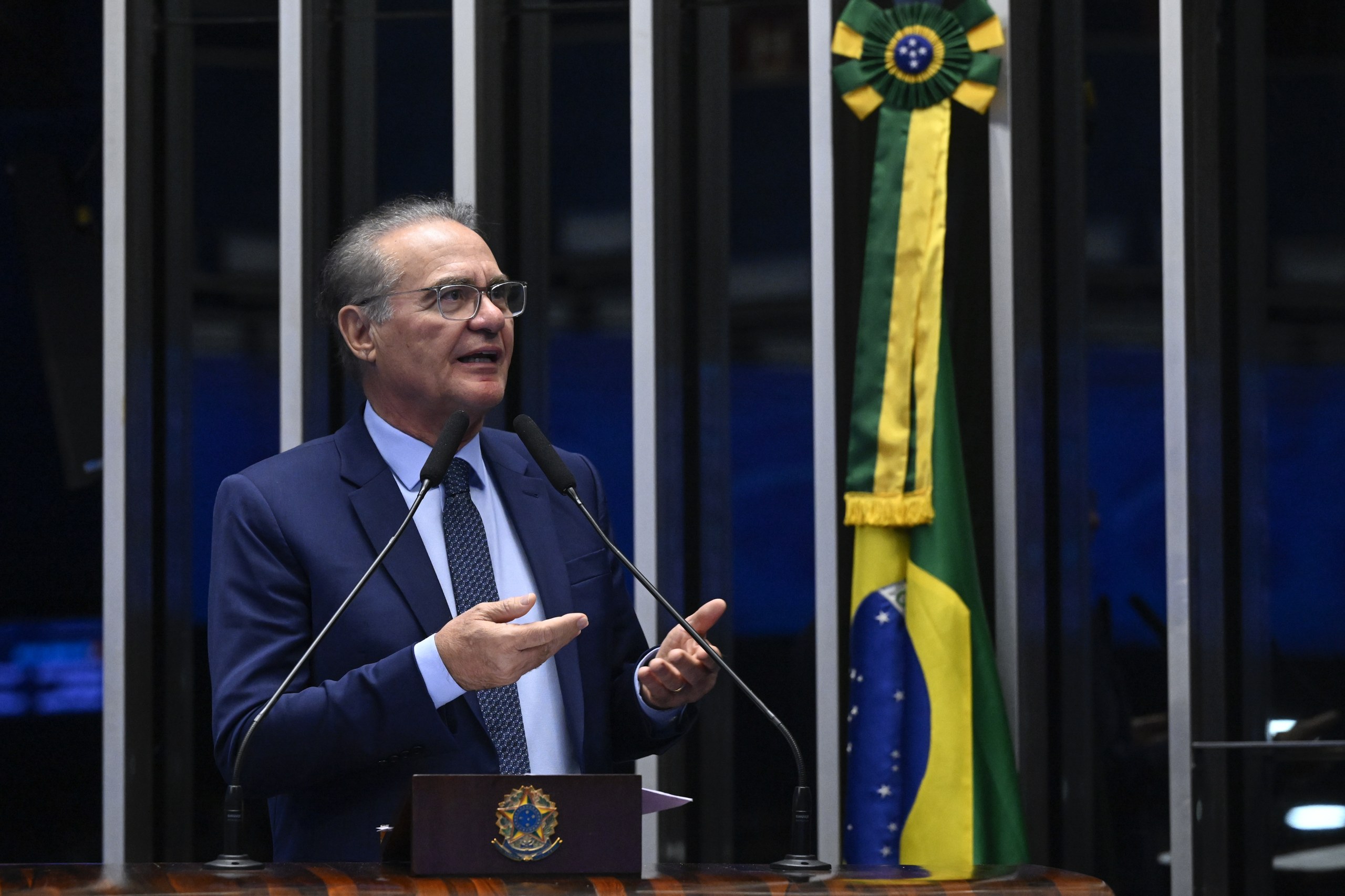 Um homem de terno e óculos discursa ao microfone em uma tribuna, com a bandeira do Brasil ao fundo em ambiente institucional. Na foto: Renan Calheiros.
