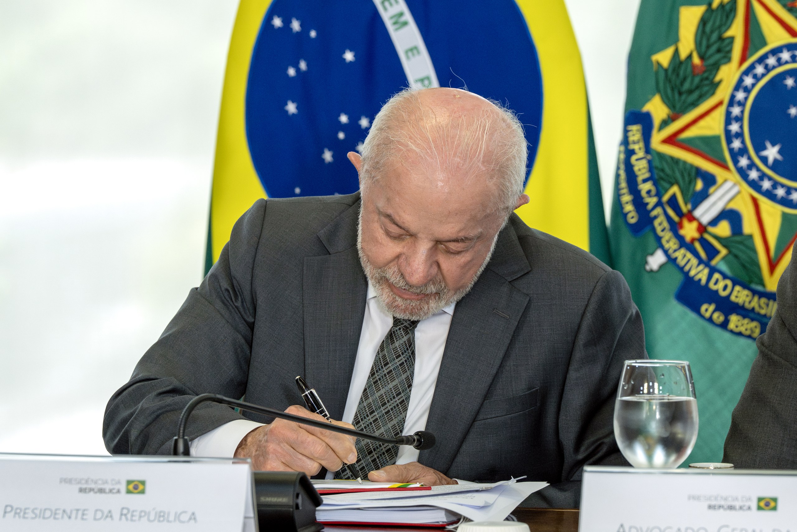 Um homem de terno e gravata assina papéis sobre uma mesa com a bandeira do Brasil ao fundo em ambiente institucional. Na foto: Luiz Inácio Lula da Silva.
