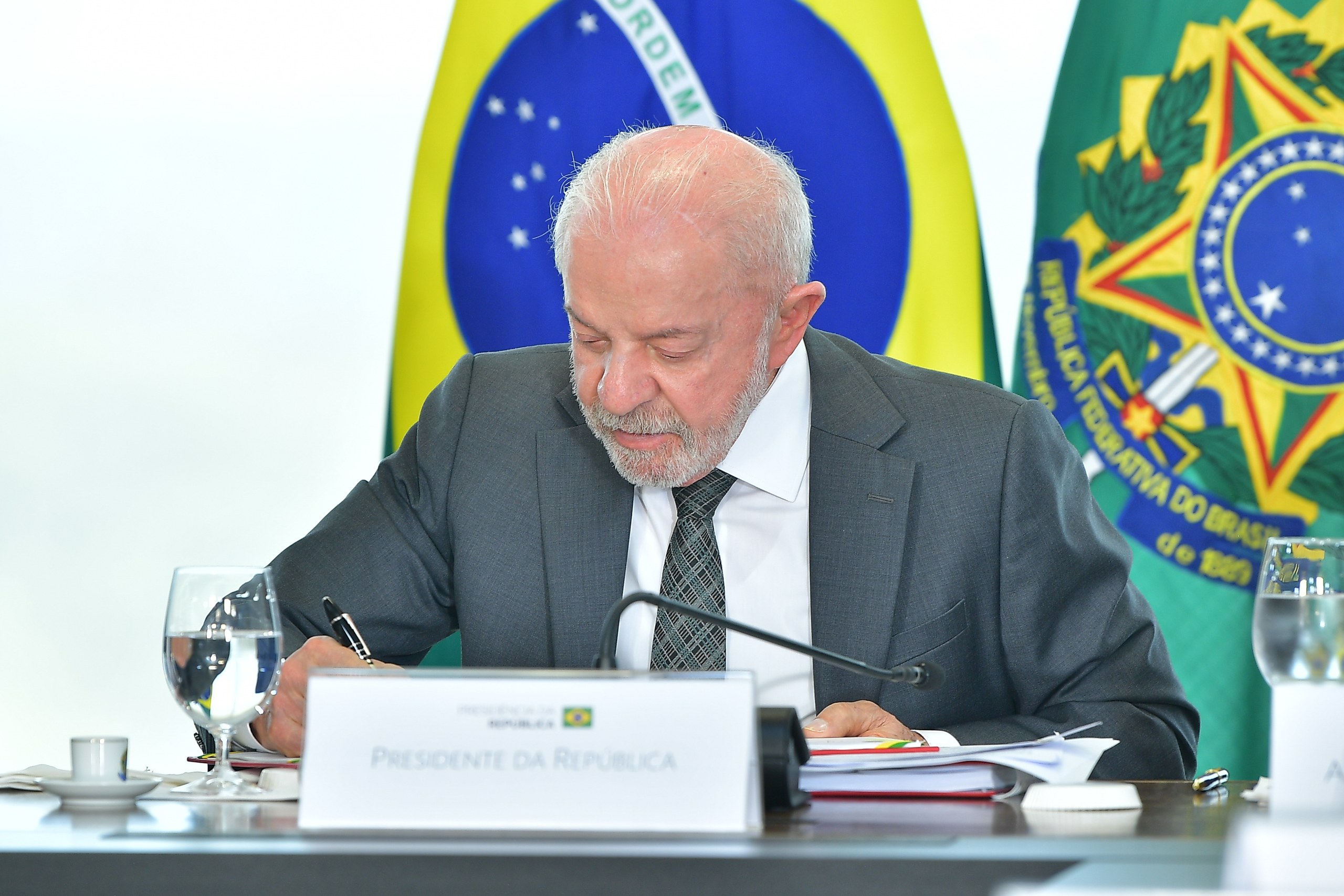 Um homem de terno e gravata assina documentos sobre uma mesa com placa de identificação, posicionado à frente de bandeiras institucionais em ambiente interno. Na foto: Luiz Inácio Lula da Silva.