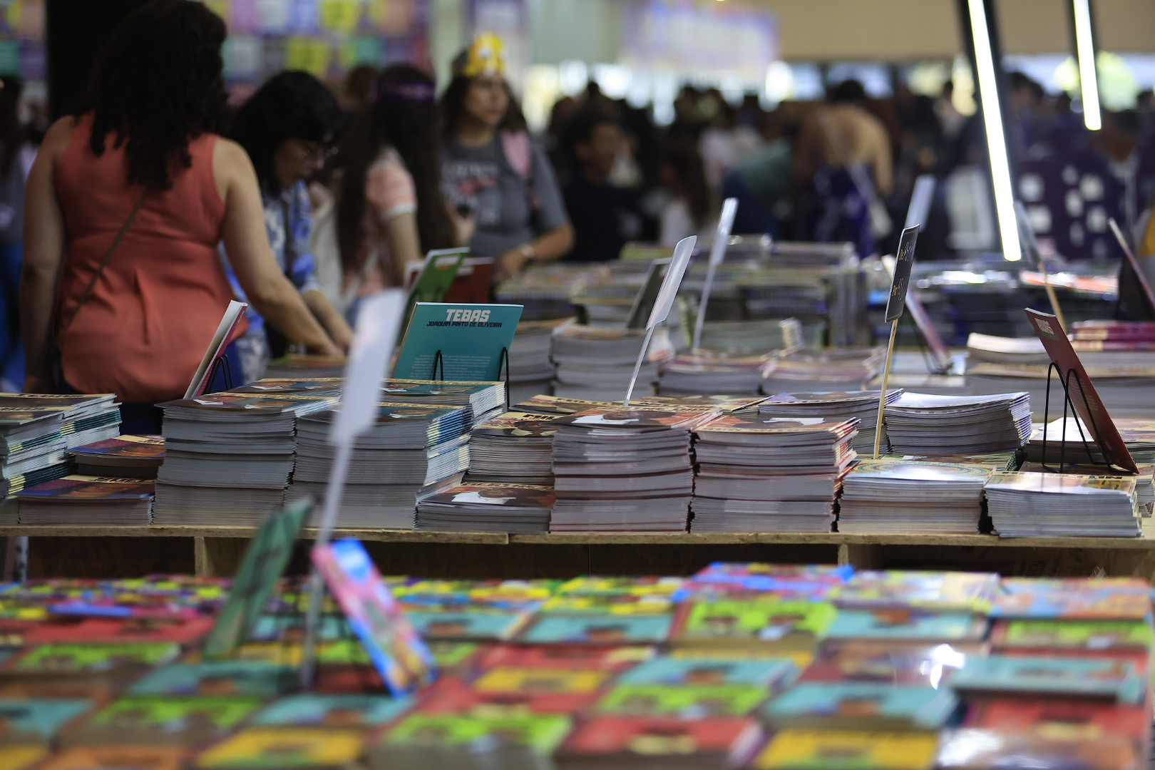 Pilhas de livros e histórias em quadrinhos organizadas sobre mesas em uma feira cultural, com pessoas circulando ao fundo.