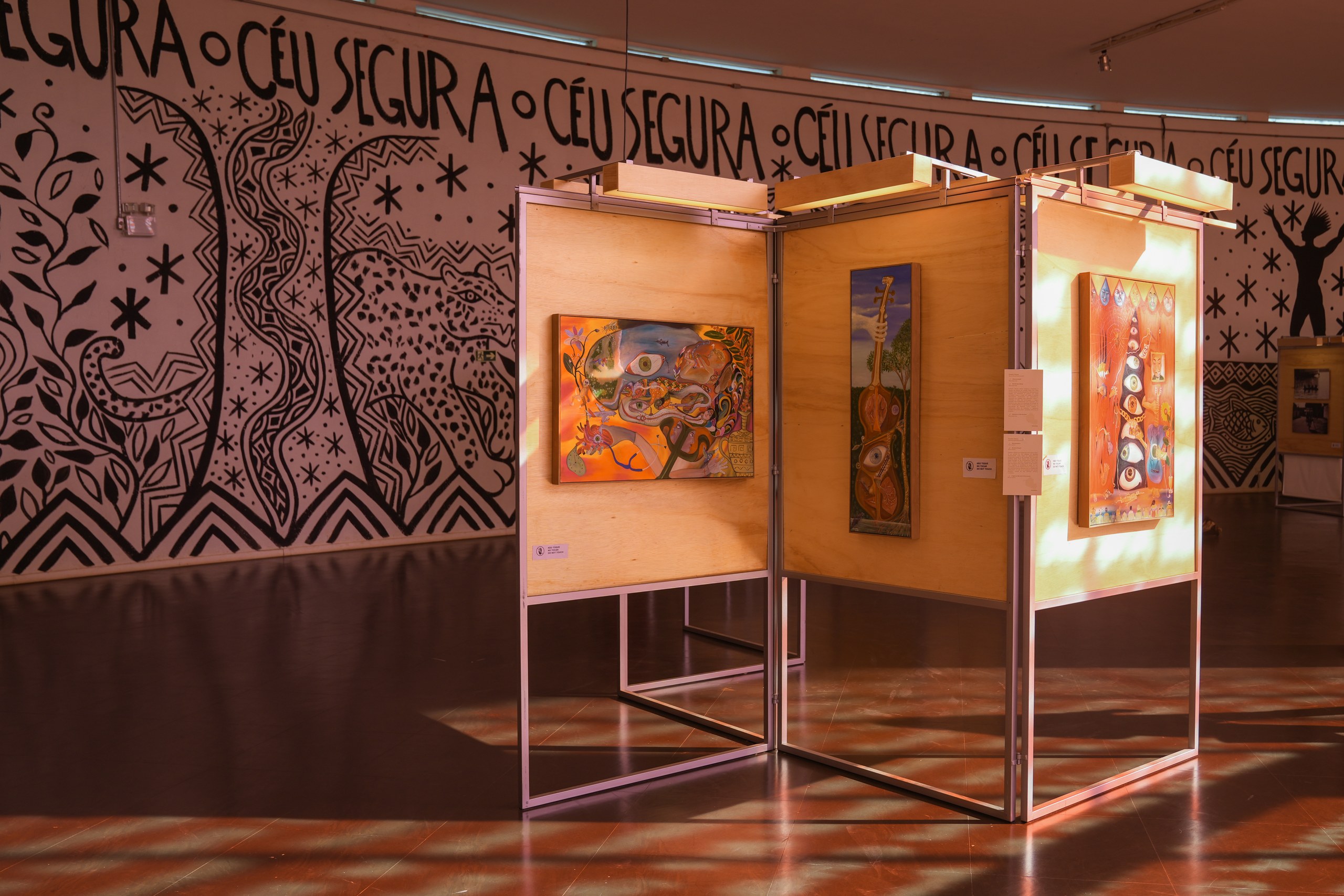 Três quadros coloridos expostos em painéis de madeira iluminados, posicionados em frente a uma parede branca com grafismos pretos em uma galeria de arte.