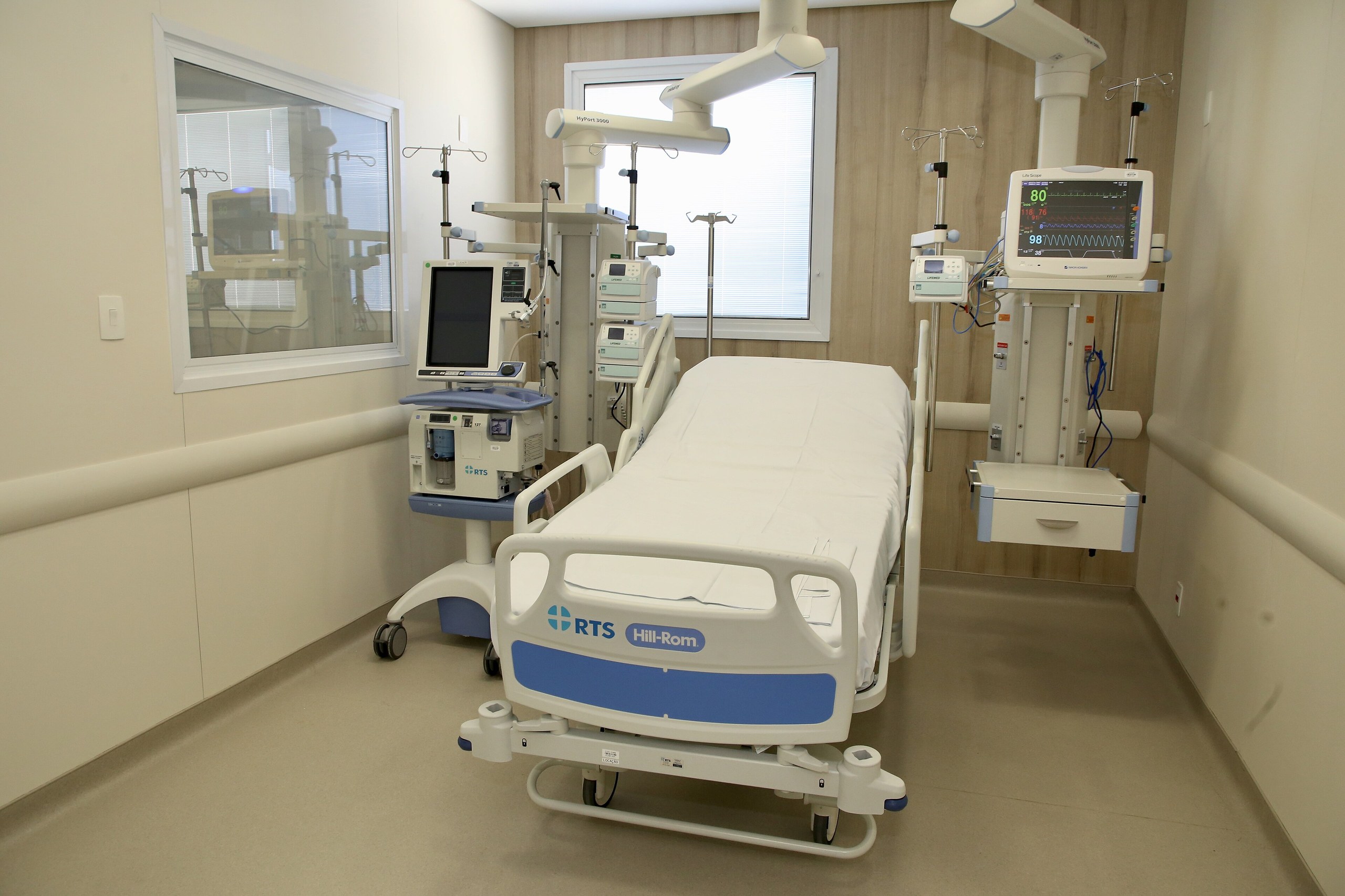 Um leito hospitalar branco com lençóis esticados, cercado por monitores e equipamentos médicos em um quarto de unidade de terapia intensiva.