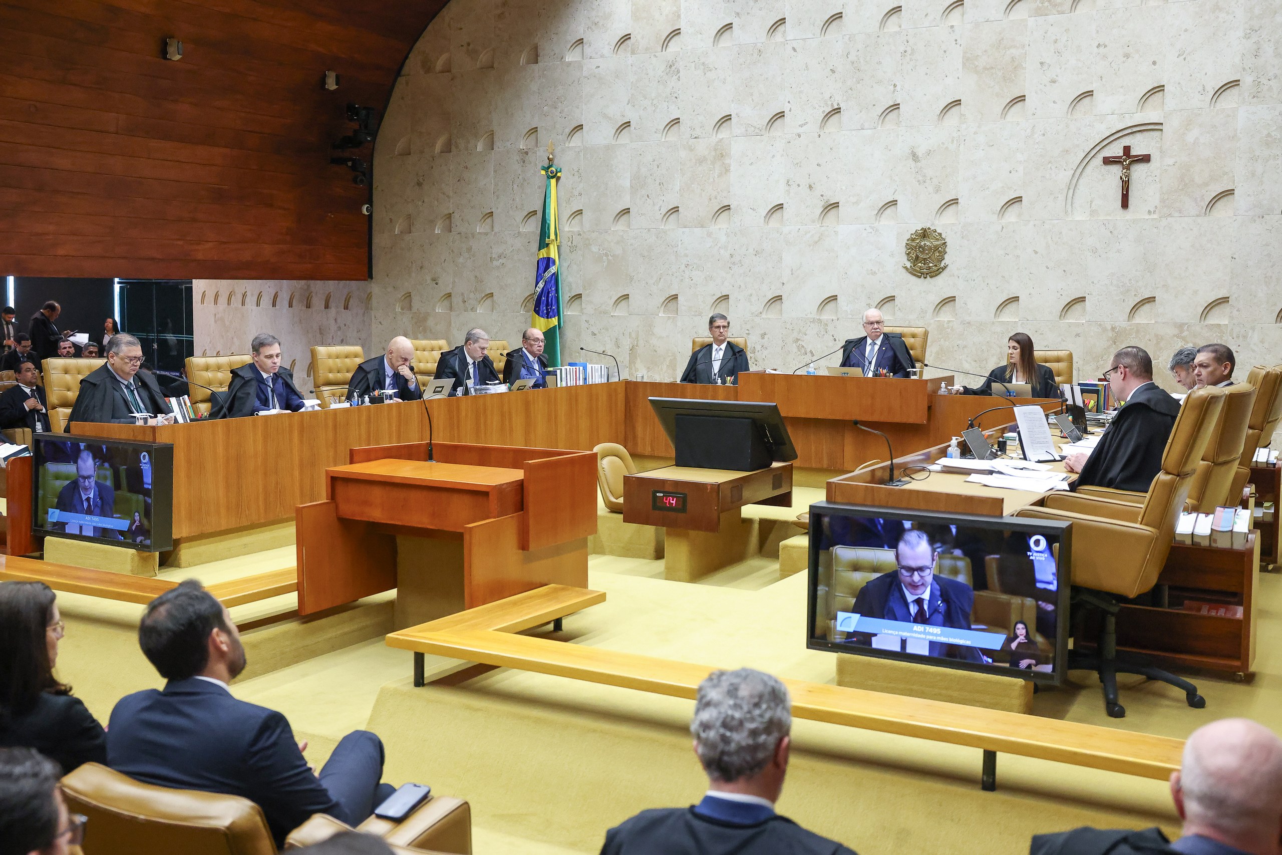 Grupo de magistrados sentados em bancada de madeira durante sessão em plenário, com monitores exibindo imagens de participantes em ambiente institucional.