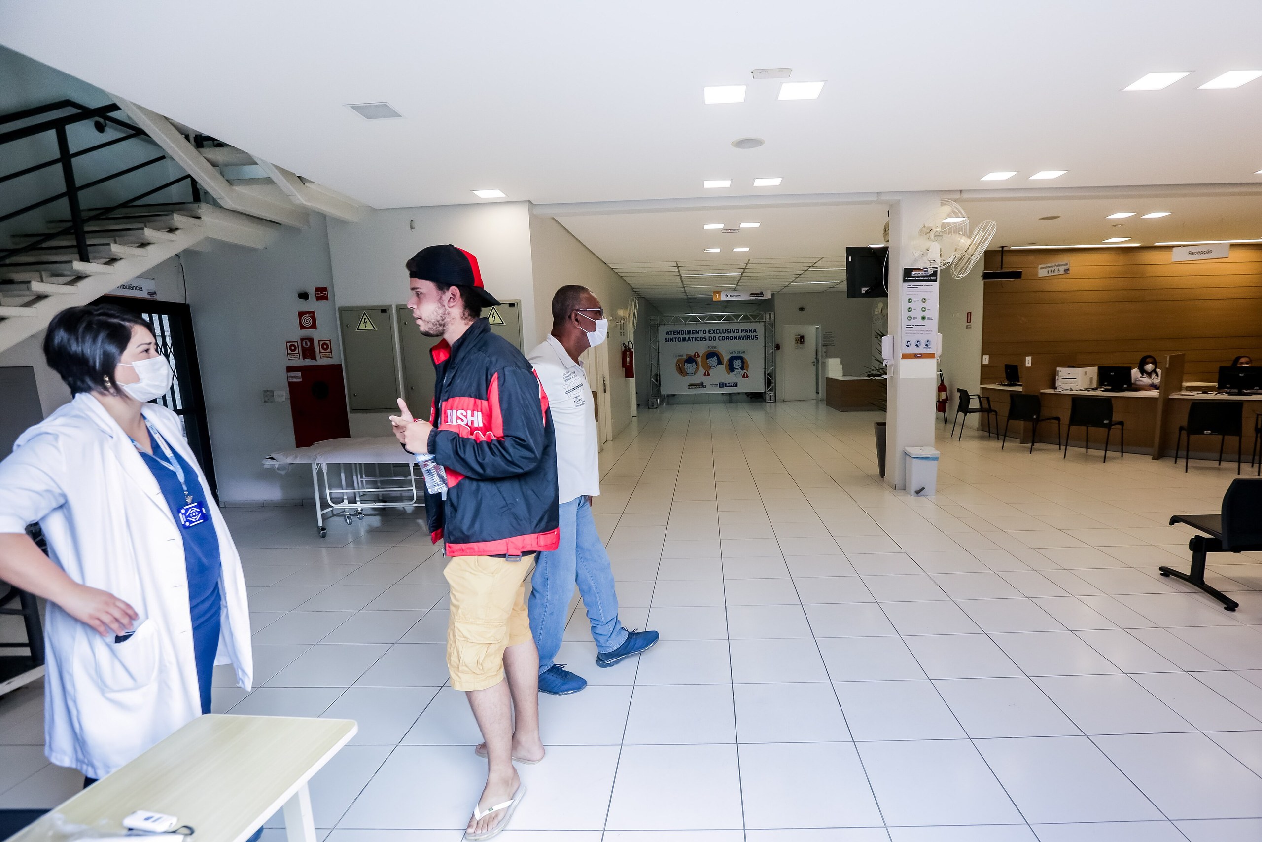 Uma profissional de saúde com jaleco e máscara conversa com um homem jovem em uma sala de espera de unidade de saúde.