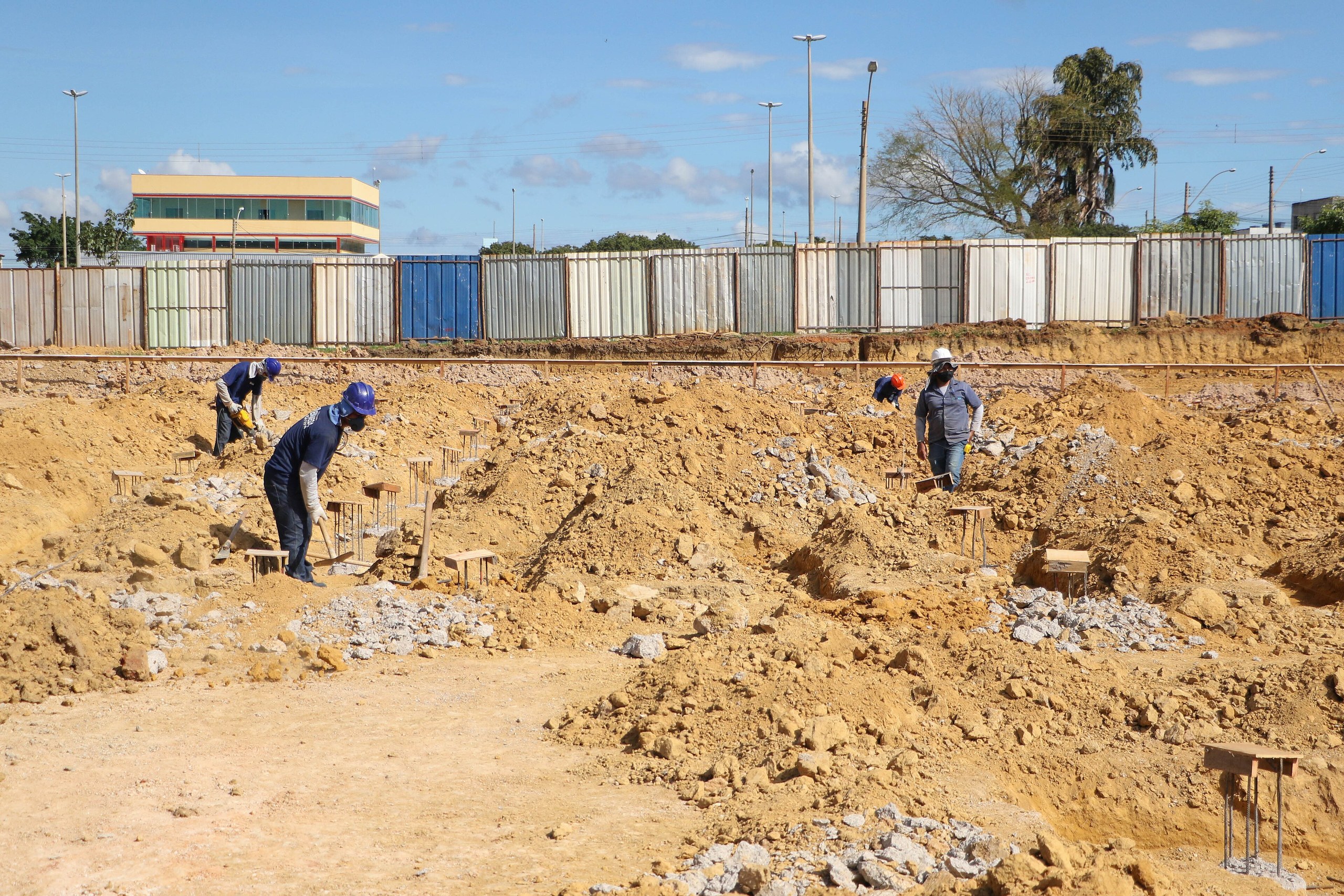 Três trabalhadores com capacetes de segurança realizam escavações em um canteiro de obras com solo arenoso sob céu azul.