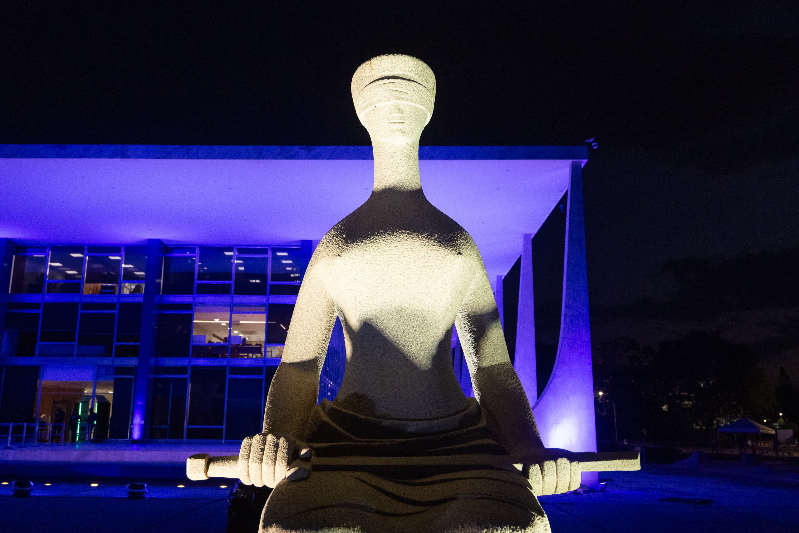 Escultura antropomórfica de pedra iluminada frontalmente, posicionada diante de um edifício moderno com luzes azuis ao fundo durante a noite.
