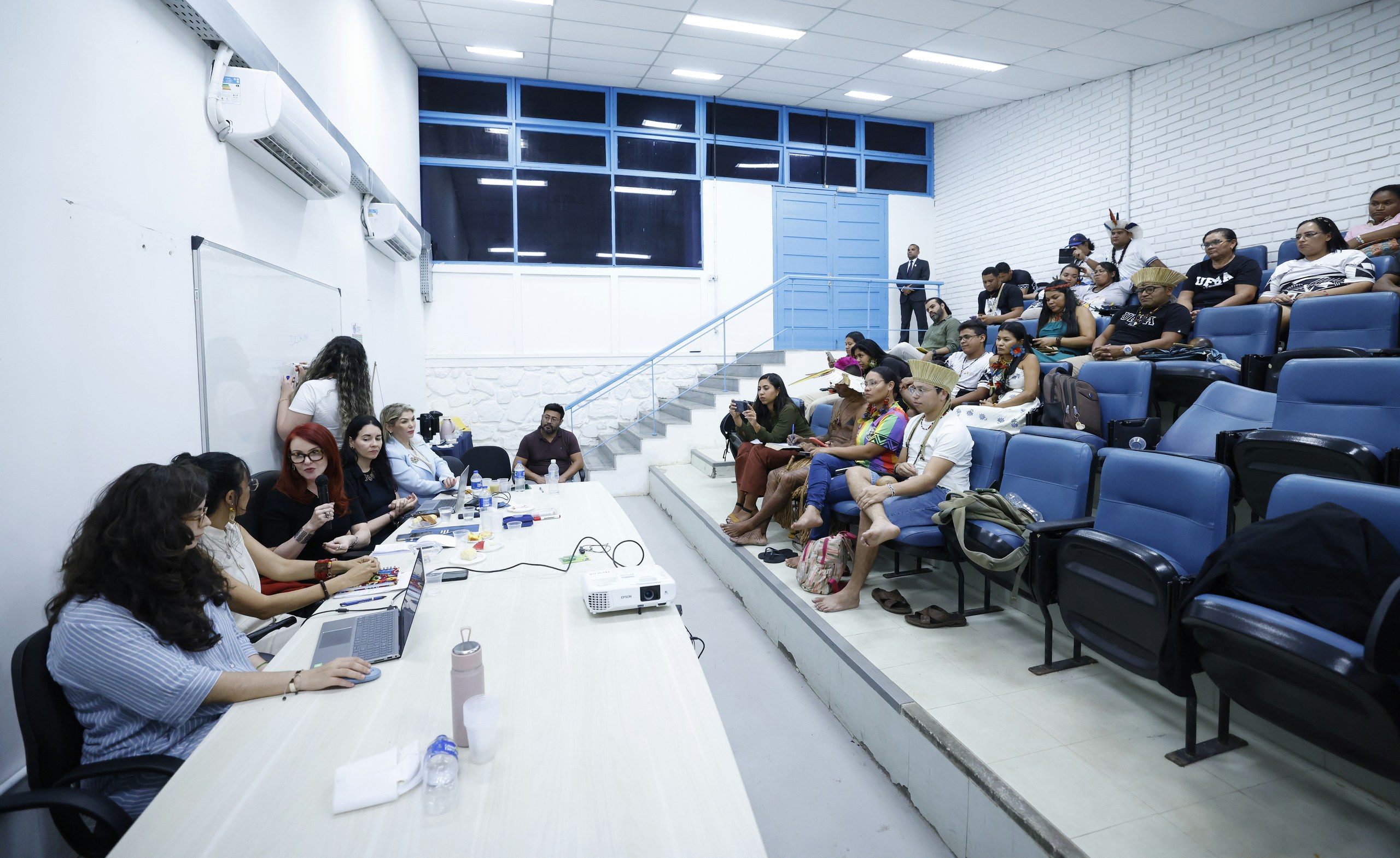 Um grupo de estudantes sentados em poltronas azuis assistem a uma aula ministrada por mulheres em uma sala com projetor e laptop.