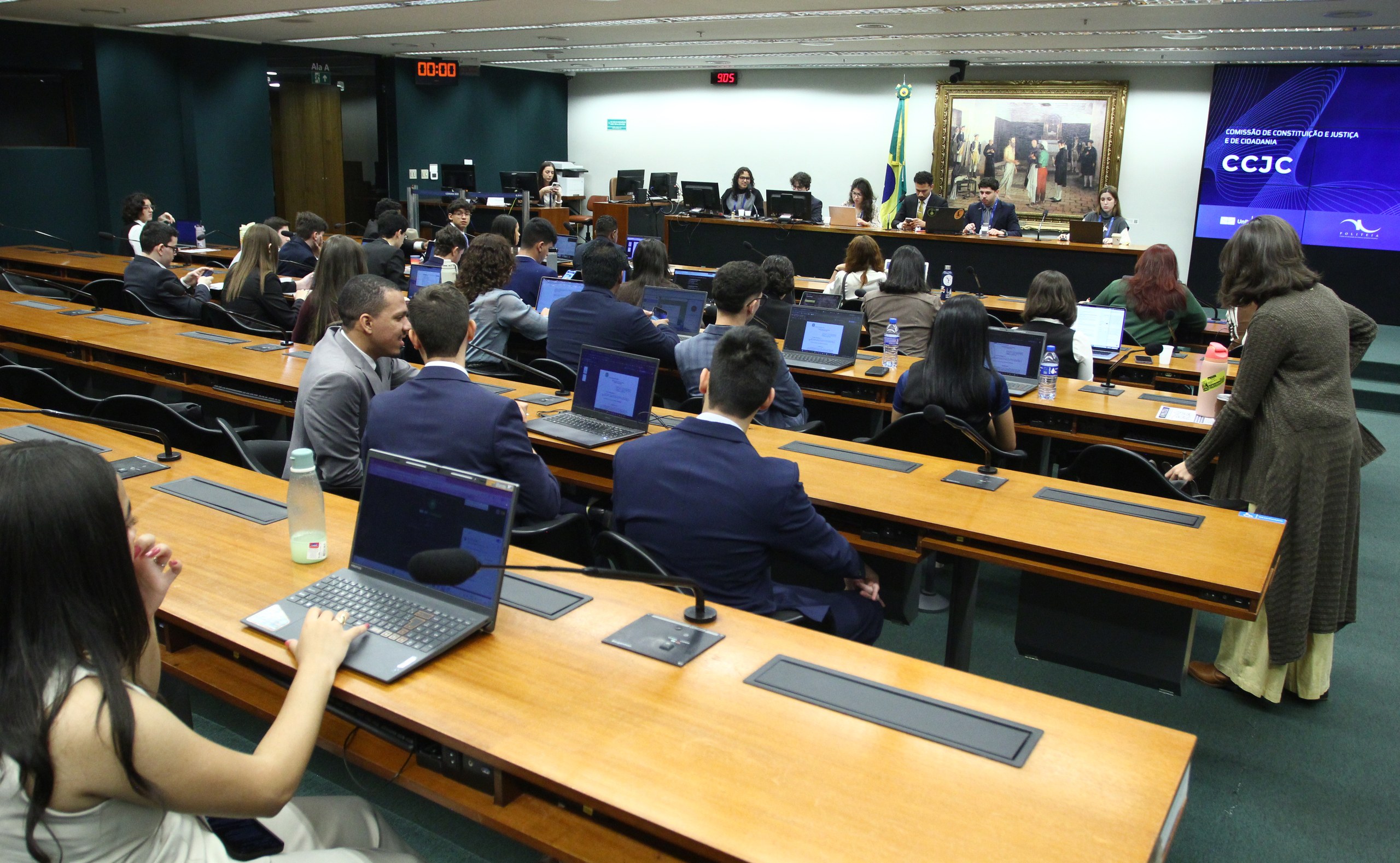 Pessoas participam de uma reunião formal em uma sala de comissões com laptops e microfones.