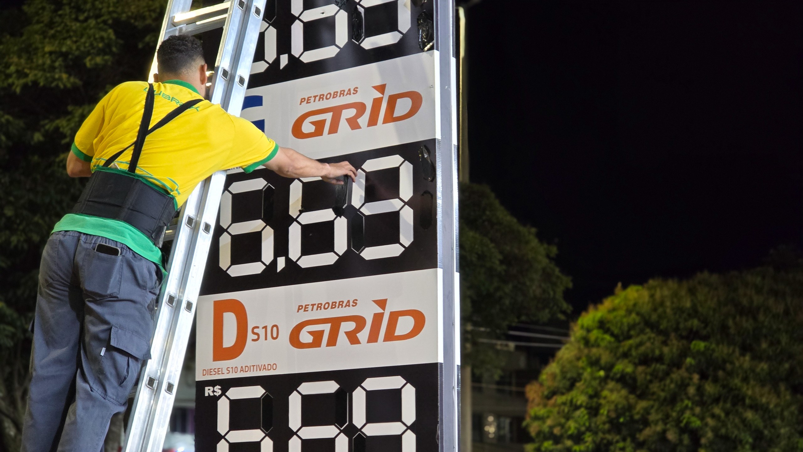 Trabalhador em uma escada realiza a manutenção de um painel de preços de combustíveis em um posto Petrobras durante a noite.