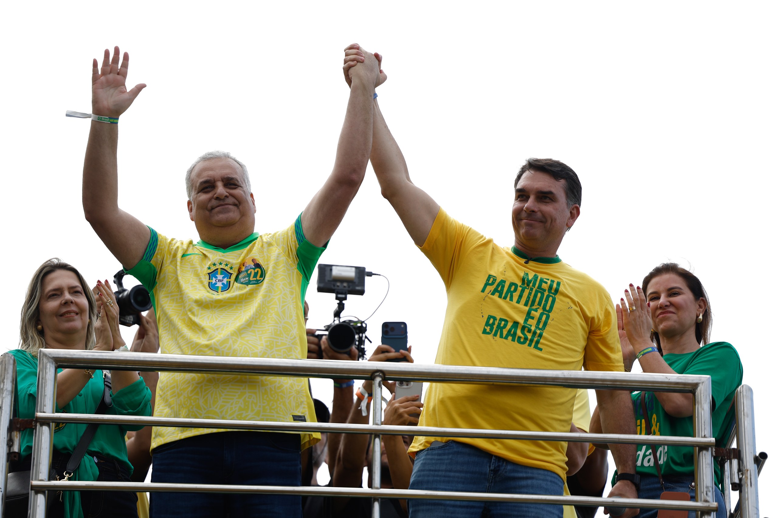 Dois homens com camisas amarelas celebram com as mãos erguidas durante um evento político ao ar livre. Na foto: Alfredo Gaspar e Flávio Bolsonaro.