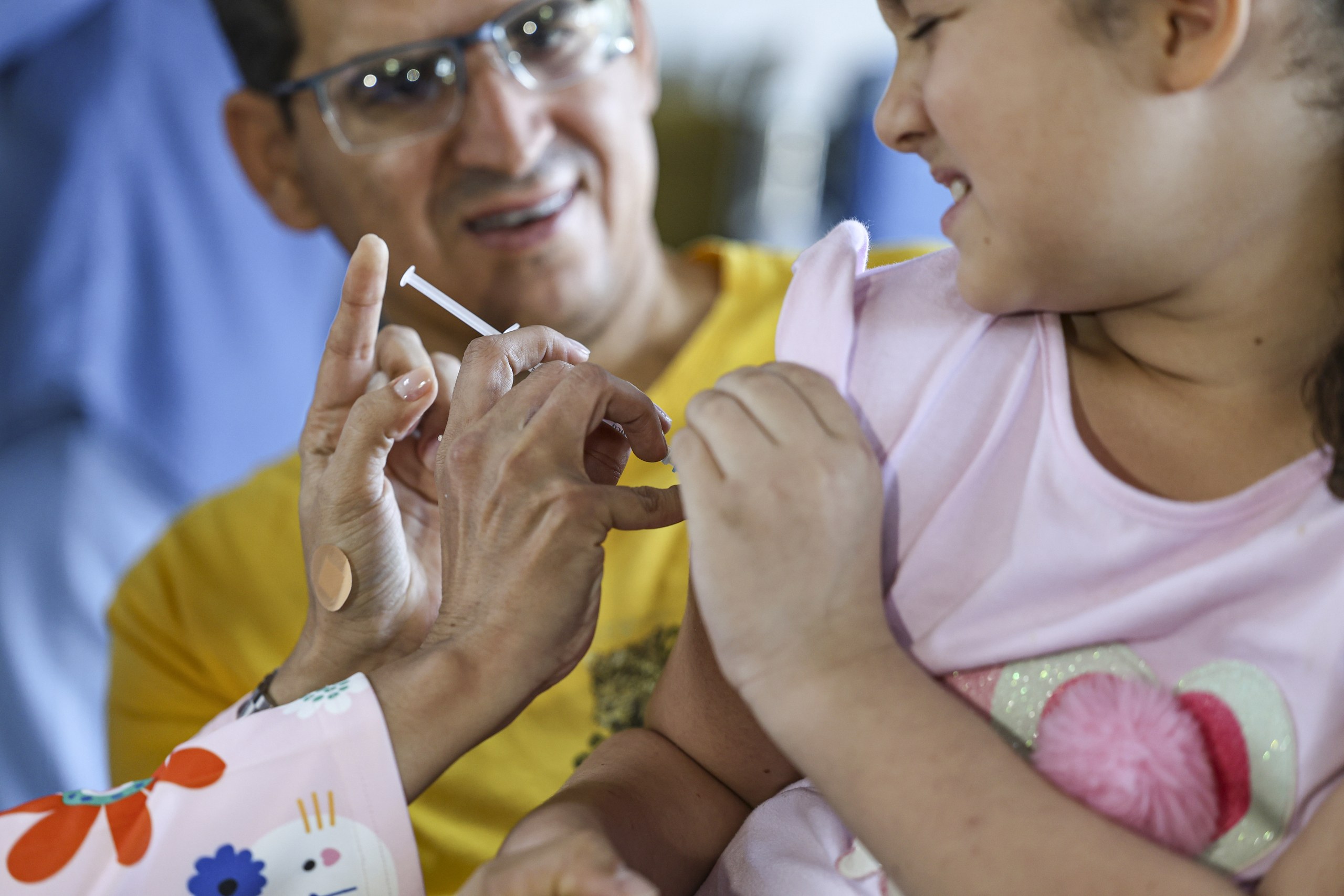 Uma criança recebe a aplicação de uma vacina enquanto é acompanhada por um adulto.