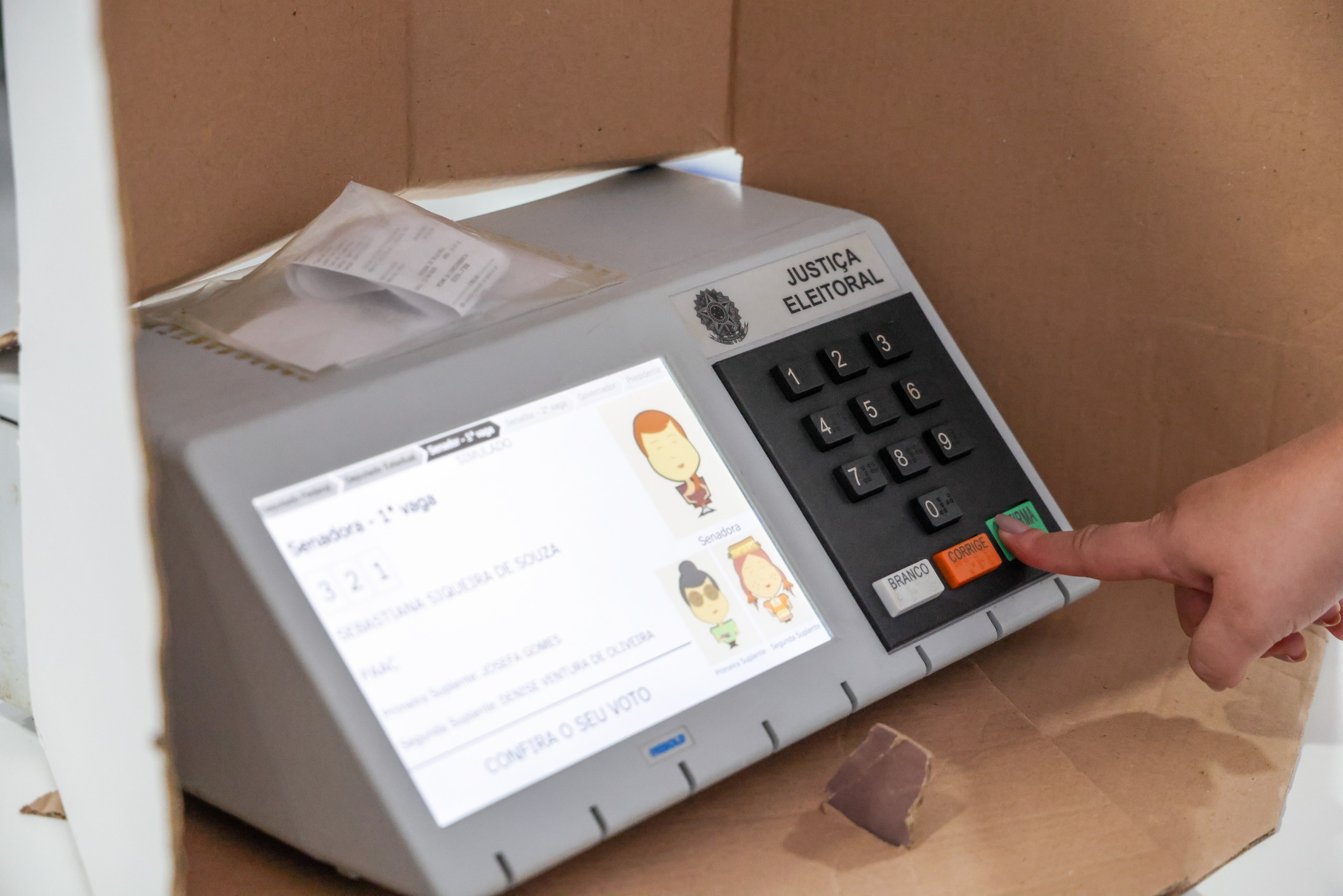 Close-up de uma mão pressionando o botão de confirmação em uma urna eletrônica da Justiça Eleitoral durante uma votação.