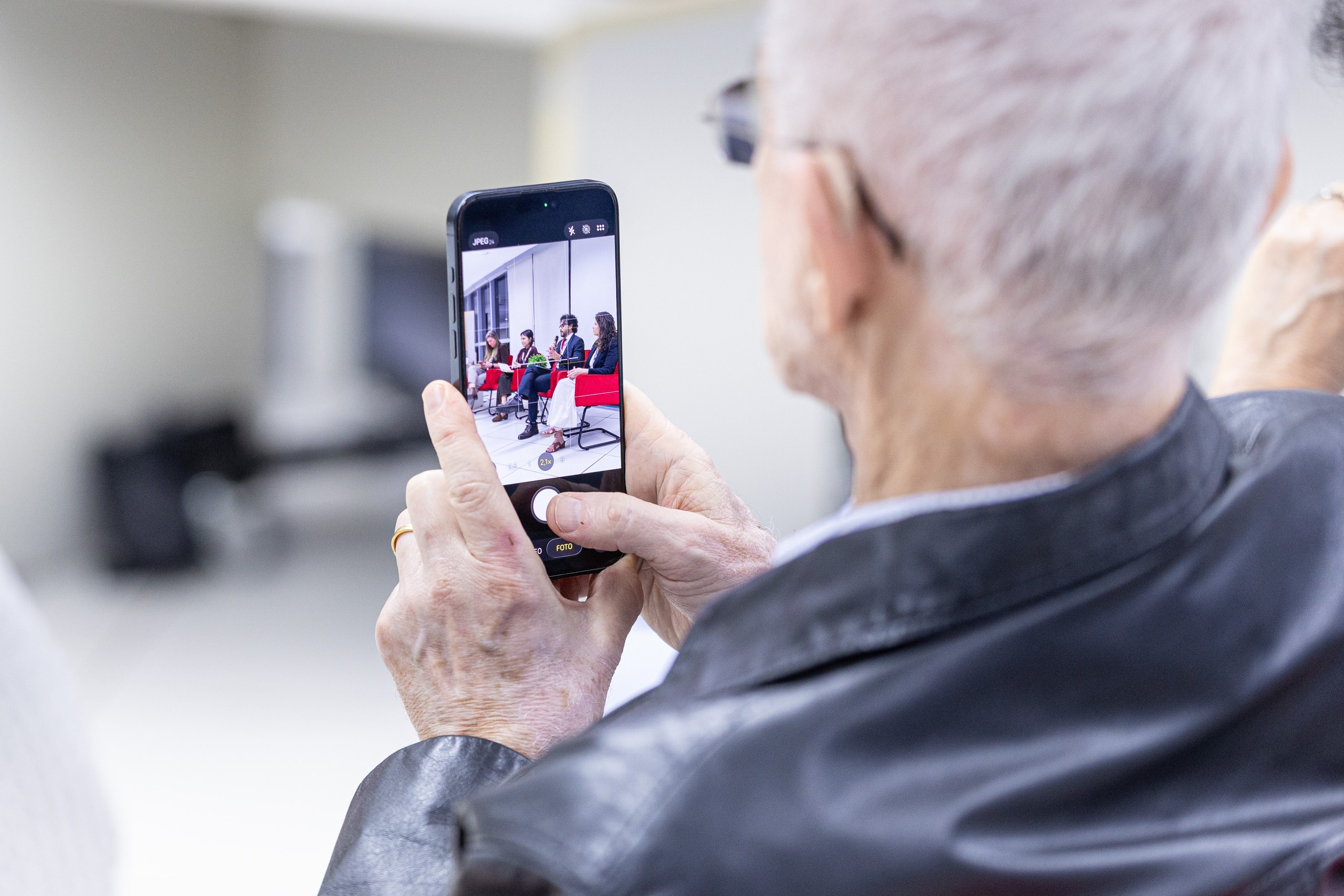 Pessoa idosa utiliza smartphone para fotografar participantes de um evento em ambiente interno.