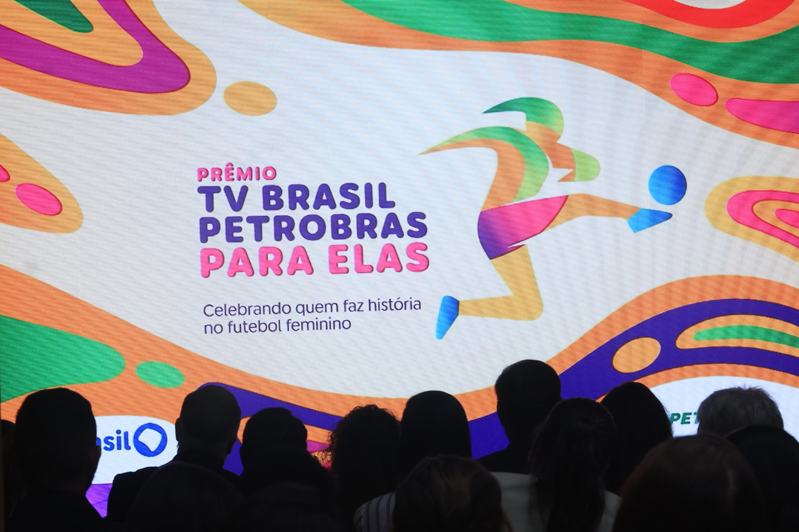Pessoas assistem a um telão anunciando o Prêmio TV Brasil Petrobras Para Elas, celebrando o futebol feminino.