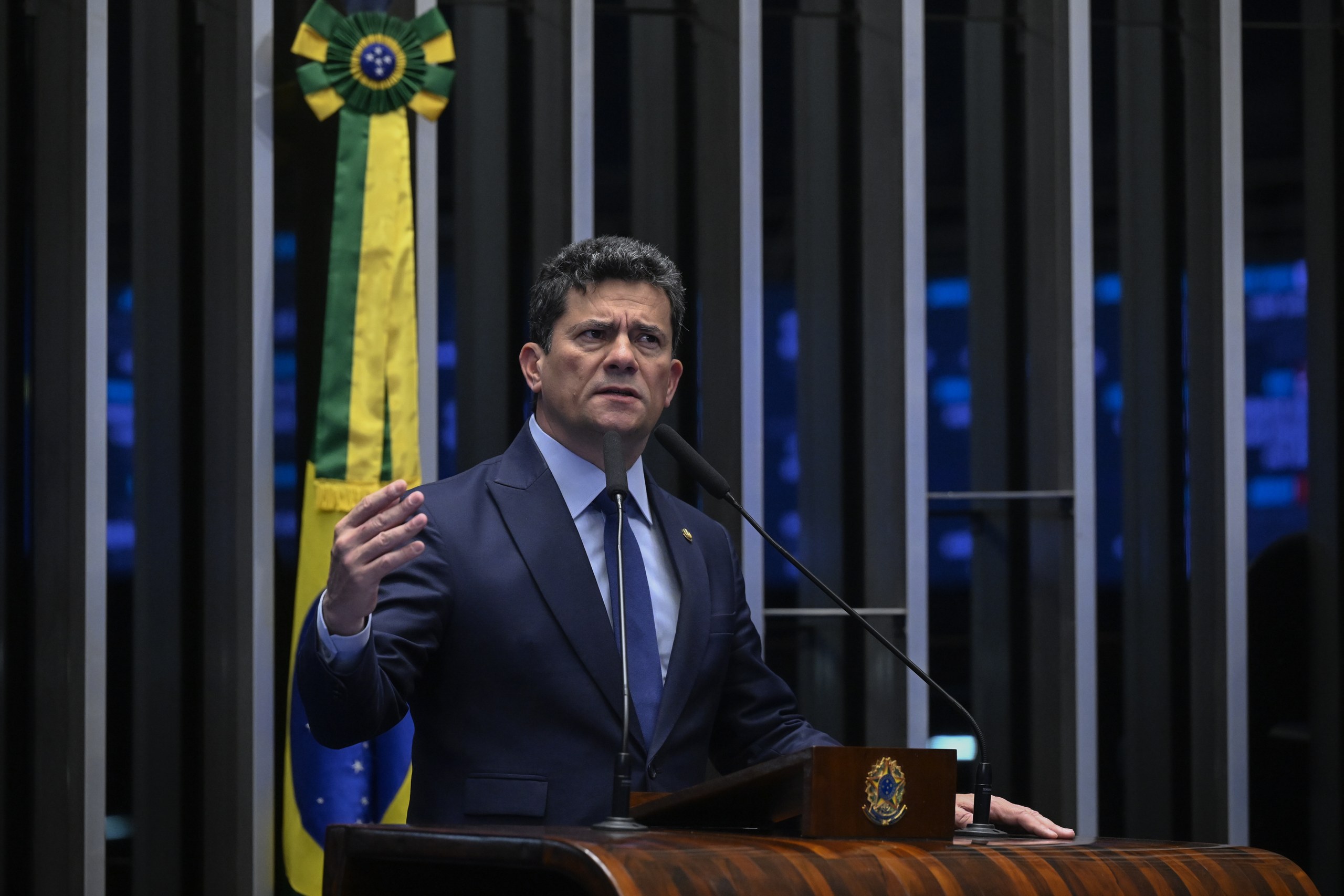 Um homem de terno e gravata discursa em um púlpito de madeira com a bandeira do Brasil ao fundo em ambiente institucional. Na foto: Sergio Moro.