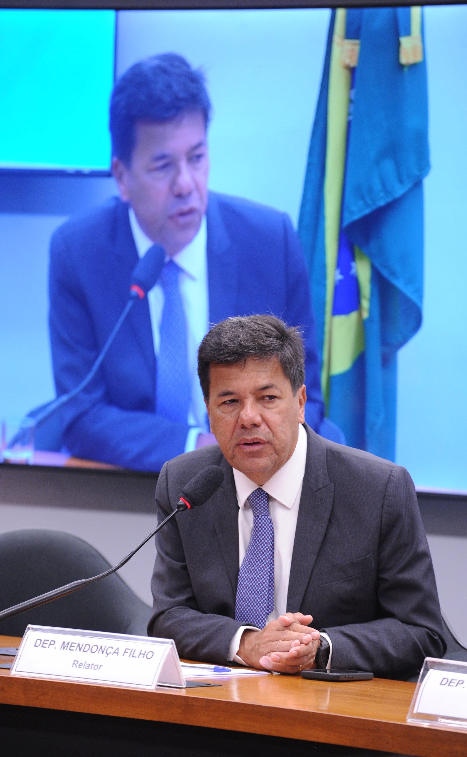 Um homem de terno e gravata sentado à mesa com microfone e placa de identificação, com sua própria imagem projetada em um telão ao fundo. Na foto: Mendonça Filho.