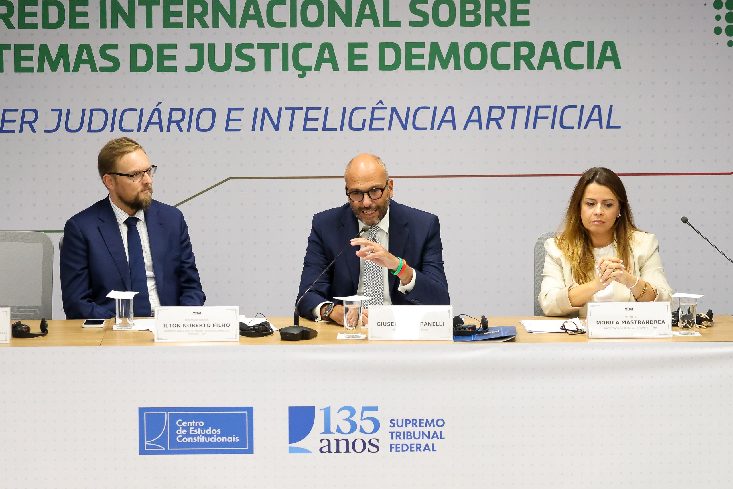 Três pessoas sentadas em uma mesa de debate com microfones diante de um painel sobre justiça, democracia e inteligência artificial em evento institucional.