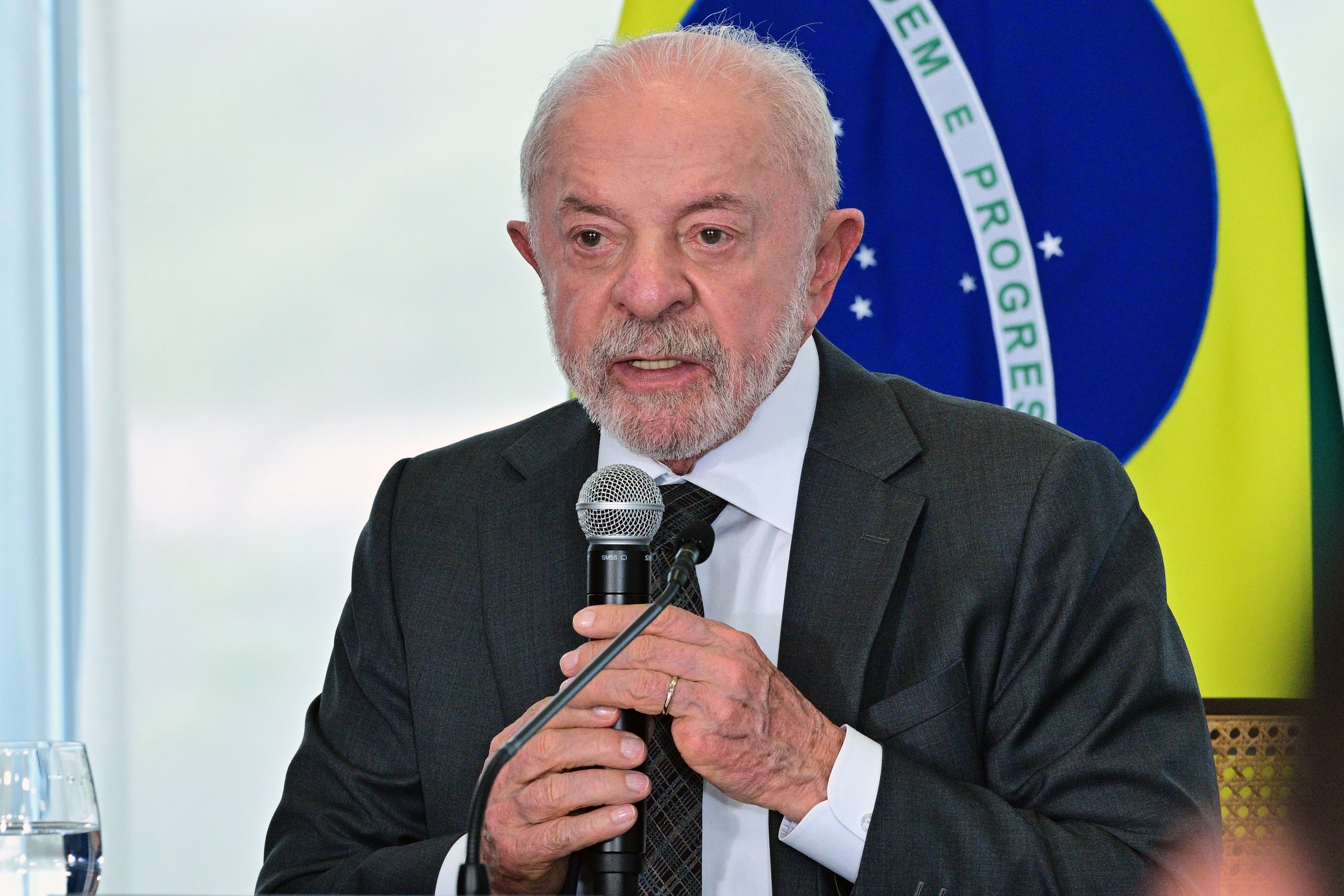 Um homem de terno e gravata fala ao microfone em frente a uma bandeira do Brasil em ambiente formal. Na foto: Luiz Inácio Lula da Silva.