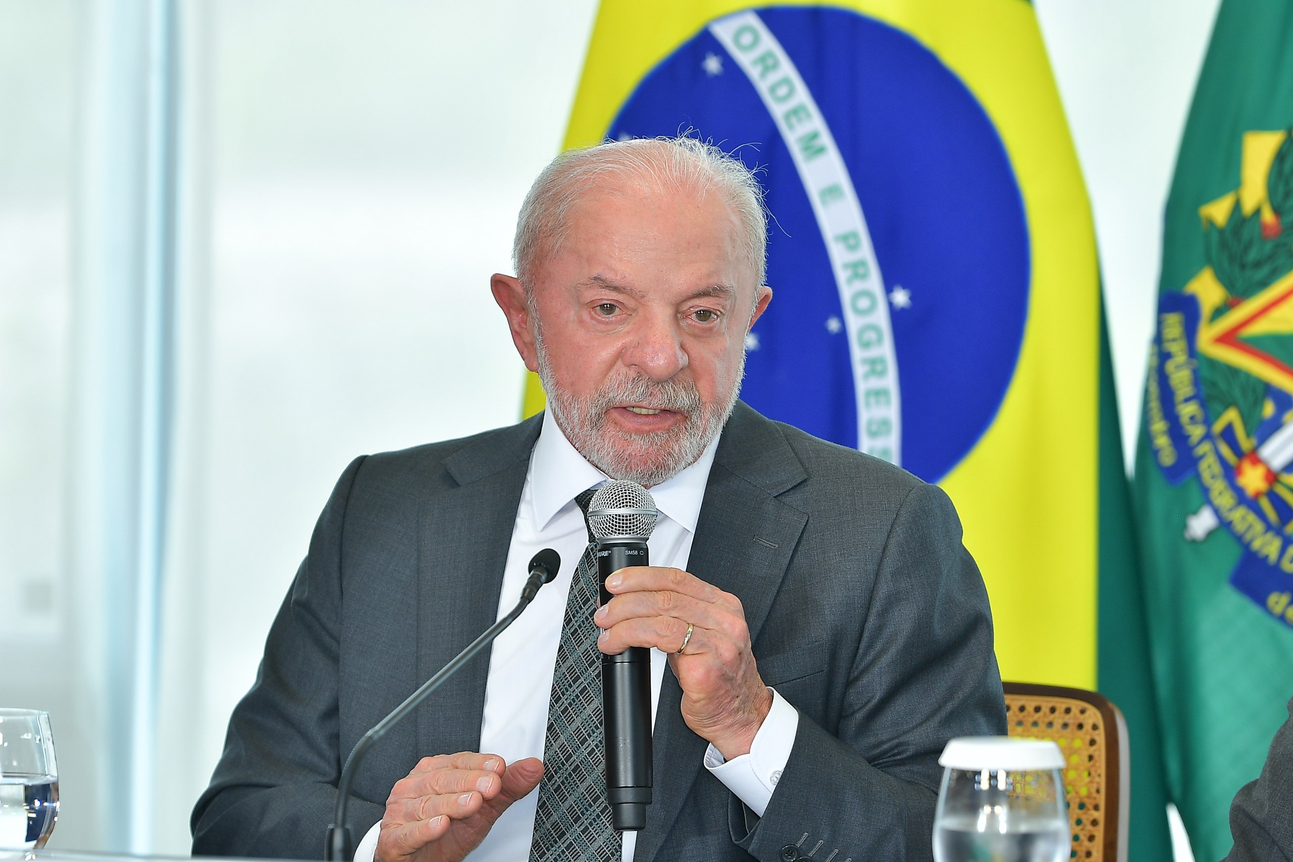Um homem de terno e gravata fala ao microfone em frente a bandeiras do Brasil e de um estado em ambiente formal. Na foto: Luiz Inácio Lula da Silva.
