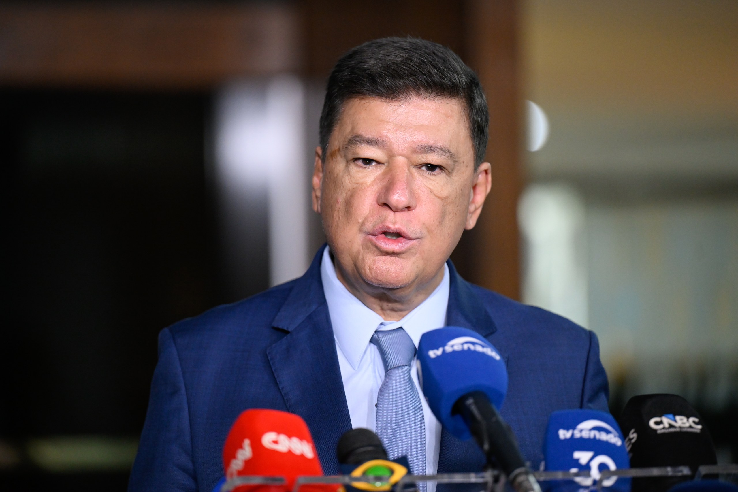Um homem de terno e gravata fala ao microfone de diversas emissoras de imprensa em ambiente interno. Na foto: Carlos Viana.
