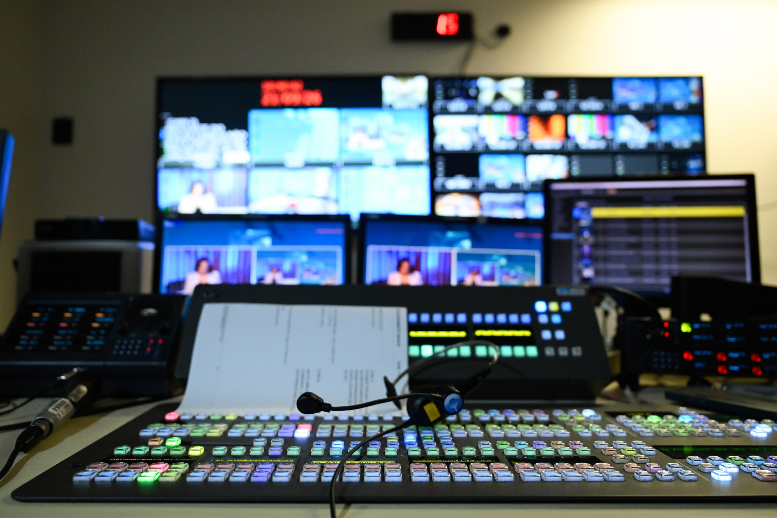 Mesa de controle de produção televisiva com botões coloridos, microfone de cabeça e monitores ao fundo exibindo diversas imagens em um estúdio.