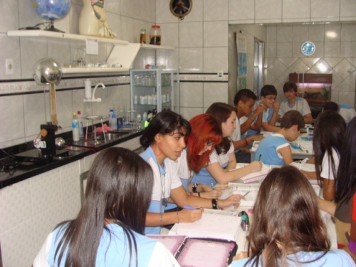 Grupo de estudantes com uniformes azuis e brancos sentados em bancadas de um laboratório escolar, escrevendo em cadernos.