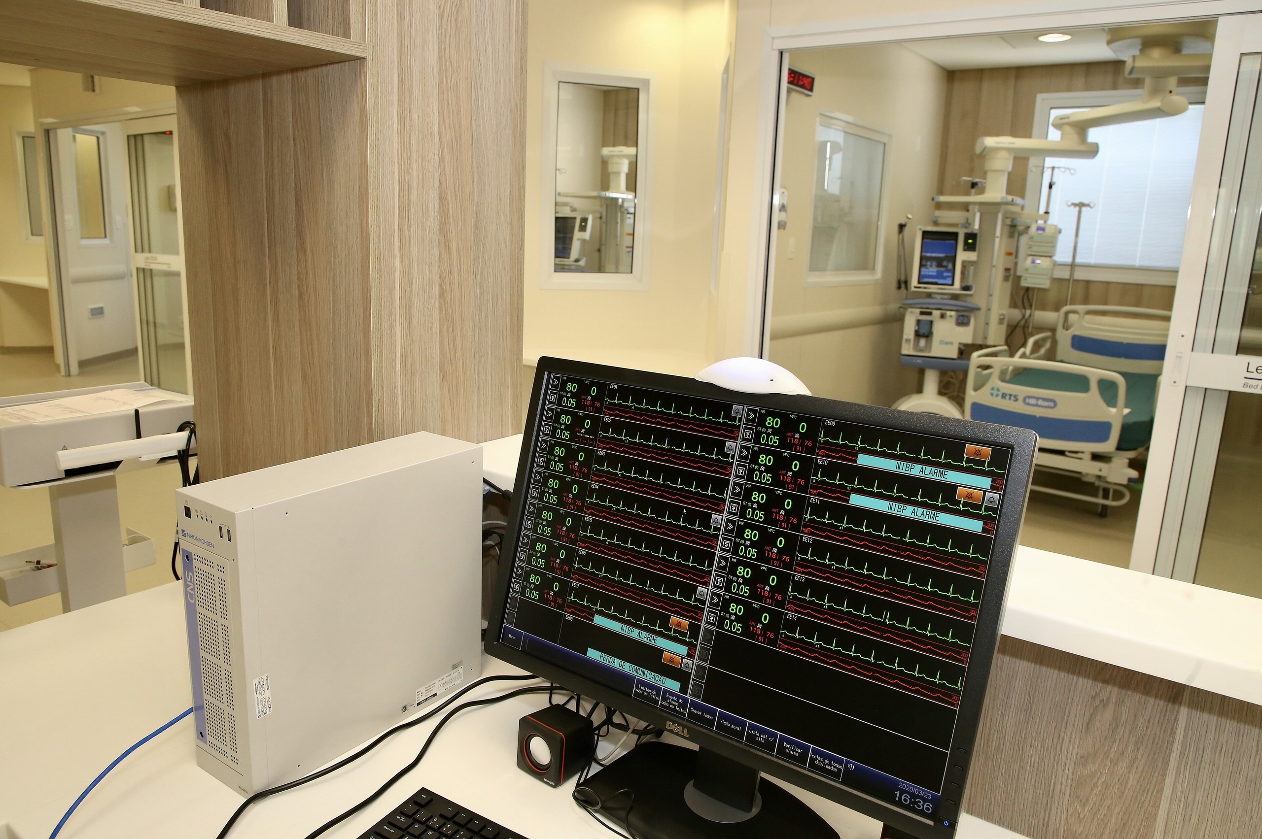 Um monitor de sinais vitais e um computador sobre uma bancada branca, com a visão de um quarto hospitalar ao fundo.