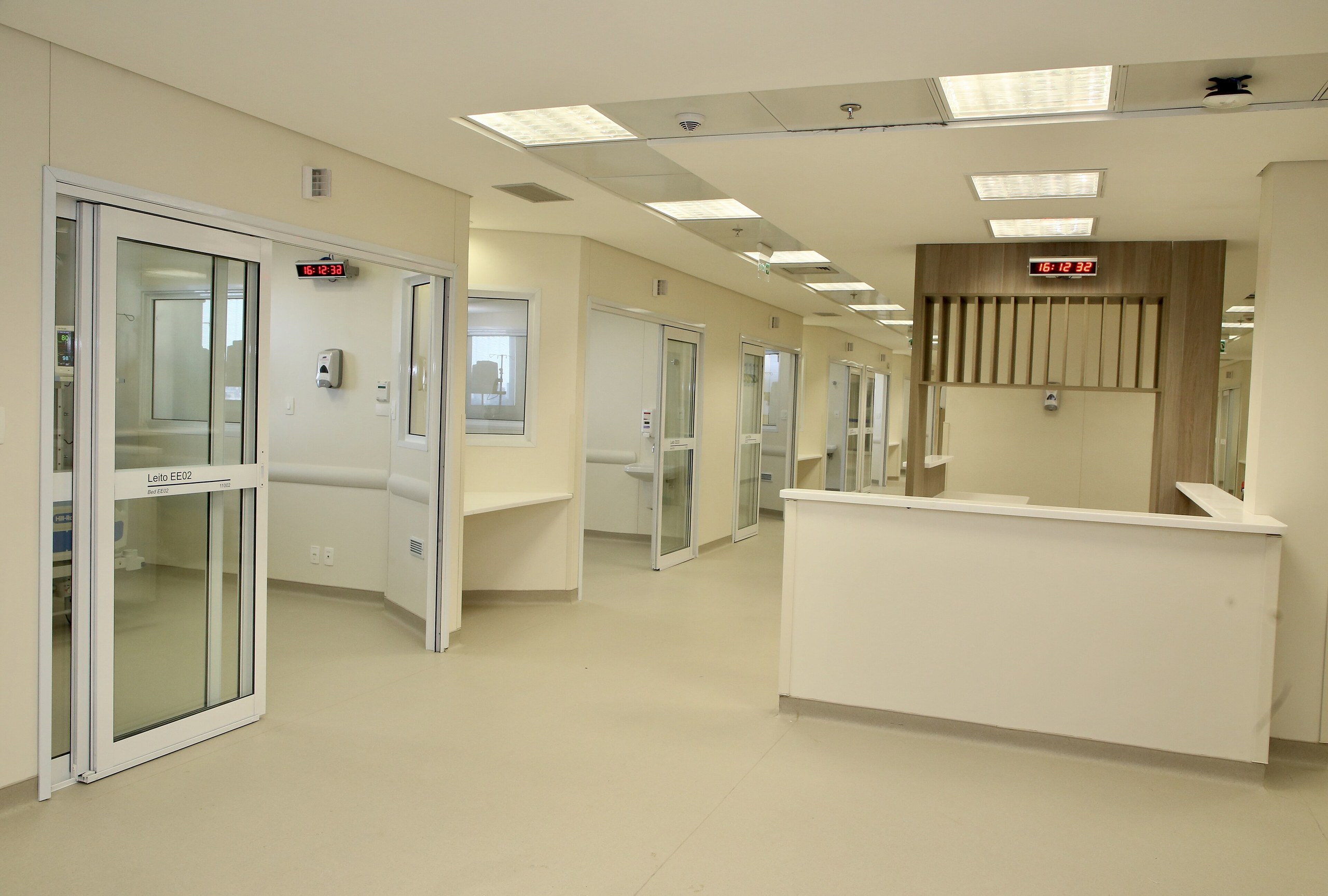Corredor de unidade de saúde com portas de vidro, balcão branco e indicação de leito em ambiente iluminado e moderno.