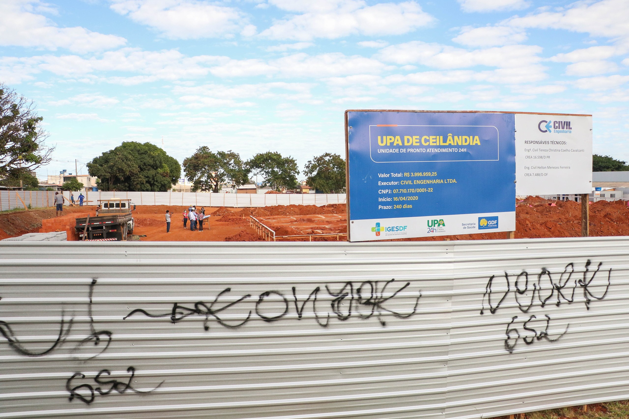 Uma placa de obra de uma Unidade de Pronto Atendimento em frente a um terreno com terra batida e operários ao fundo.