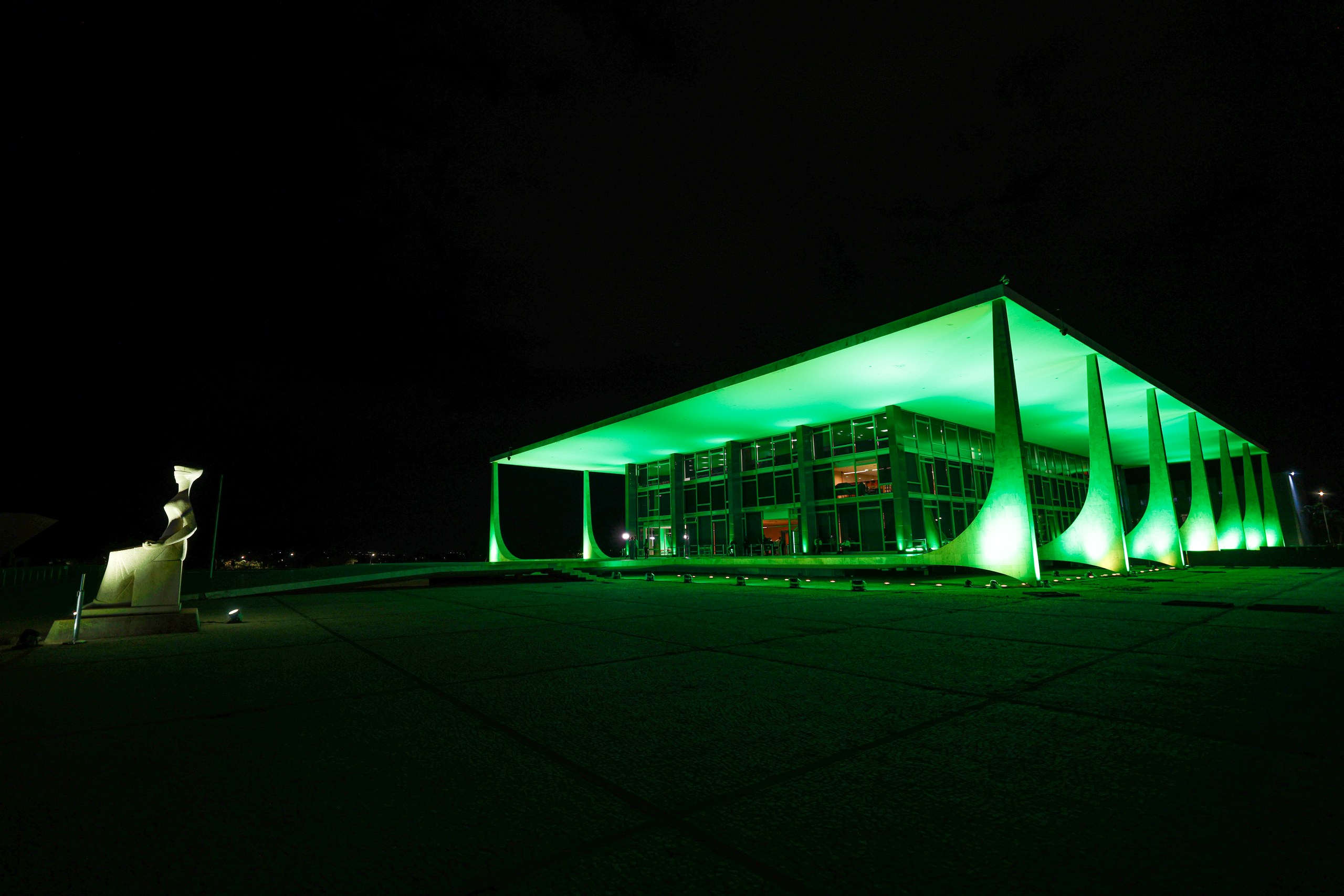 Prédio do Palácio do Planalto iluminado com luzes verdes durante a noite, com uma escultura branca ao lado em uma ampla esplanada em Brasília.