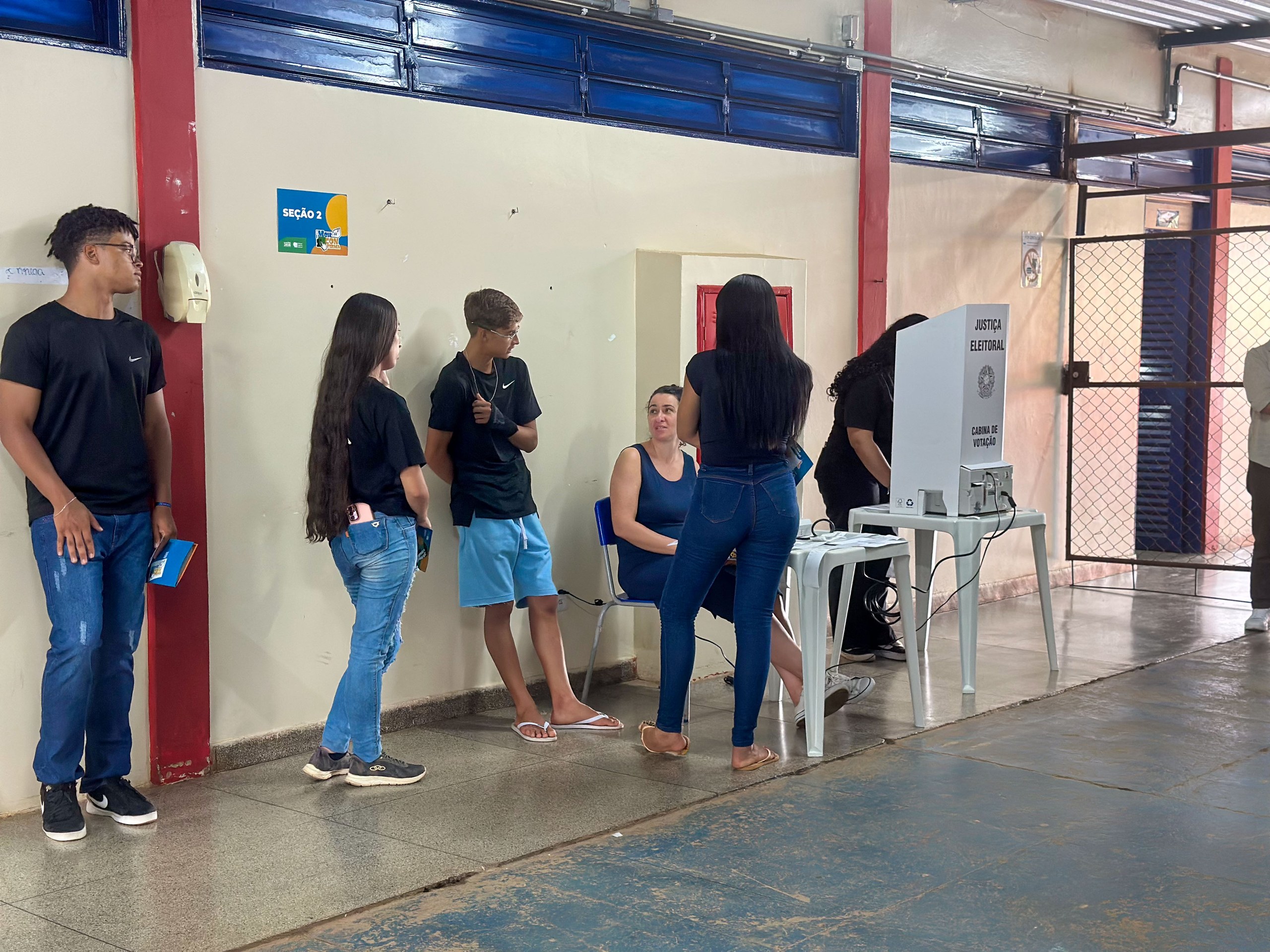 Um grupo de pessoas aguarda em fila para votar em uma urna eletrônica instalada em uma seção eleitoral dentro de um prédio público.