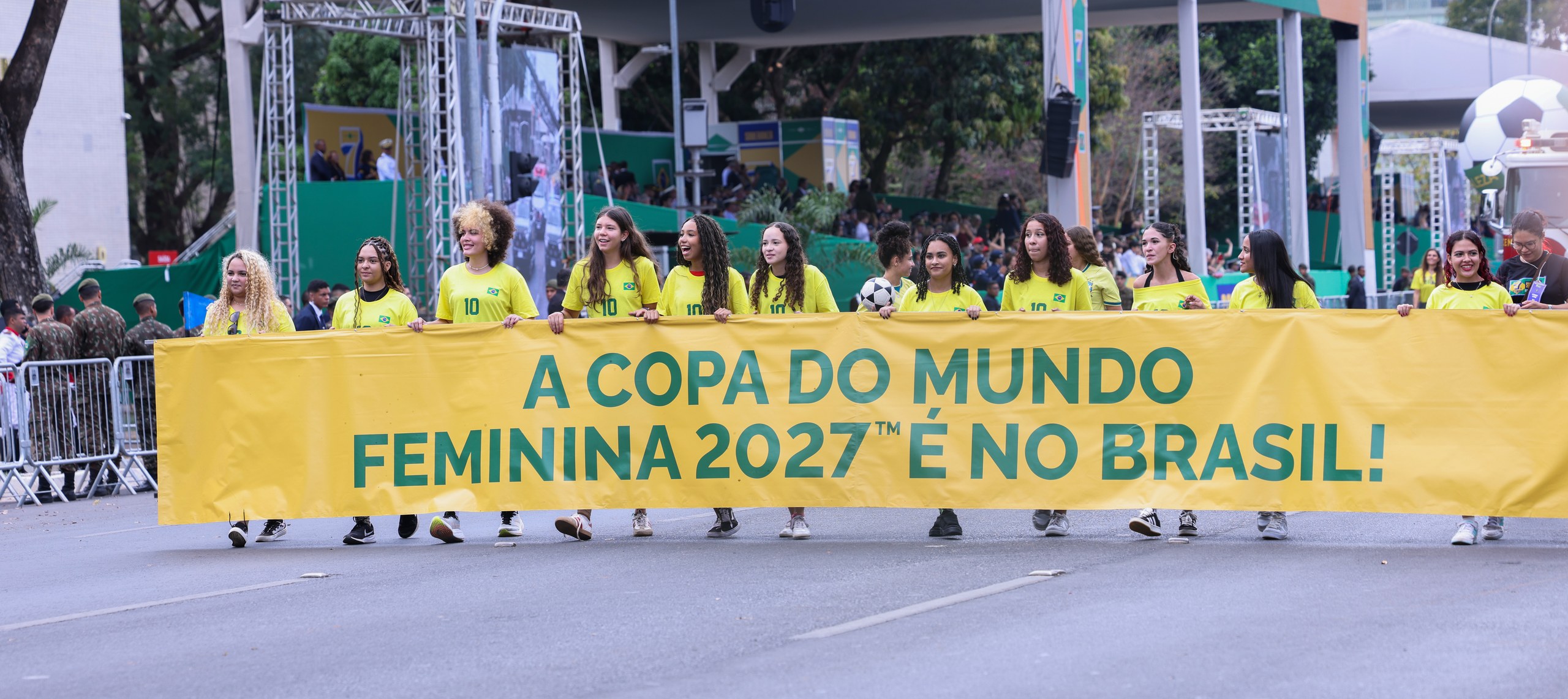 Grupo de mulheres com camisas amarelas segurando faixa amarela com texto sobre a Copa do Mundo Feminina 2027 em via pública urbana.