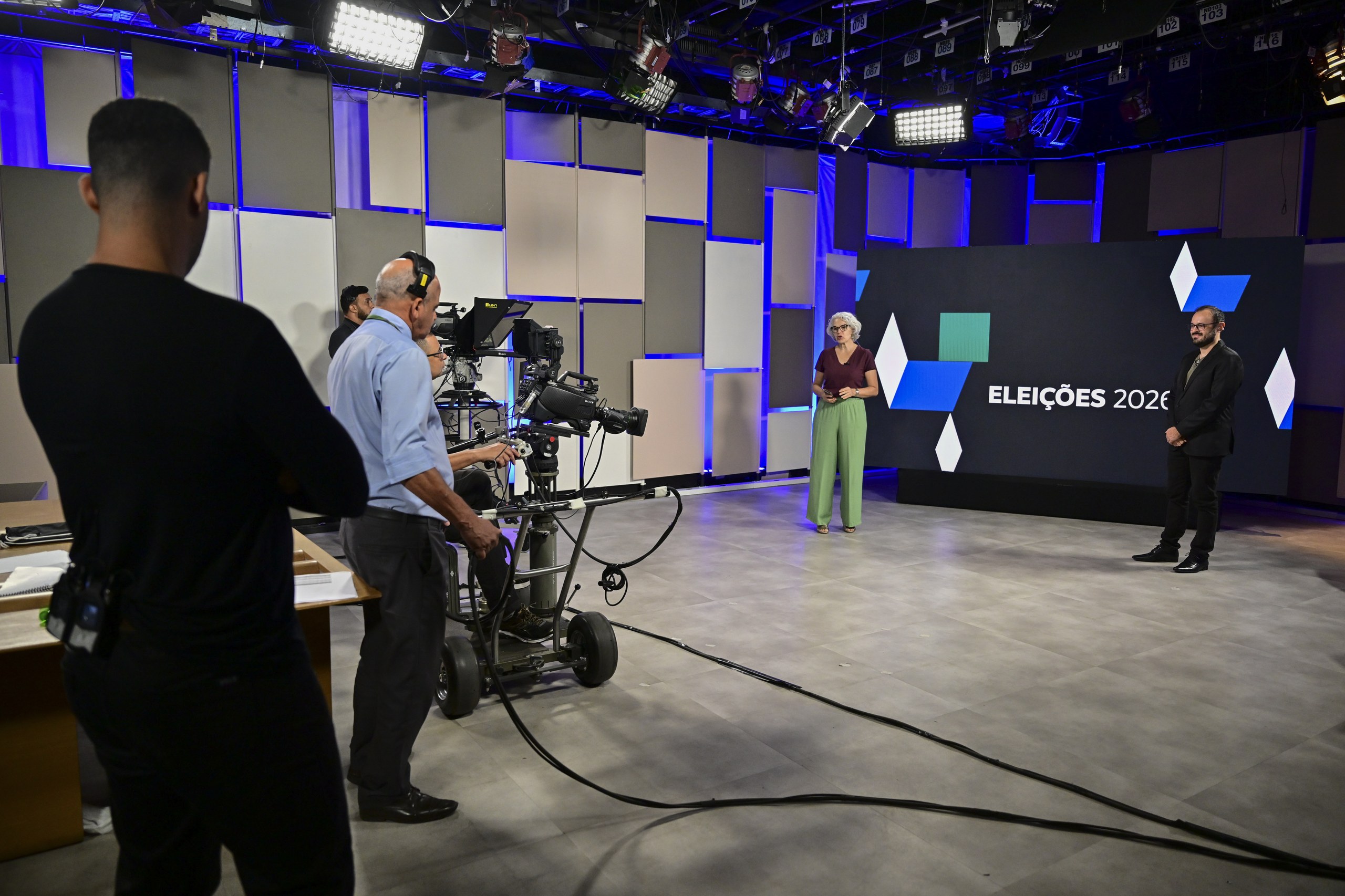 Equipe de produção opera câmera de vídeo em estúdio de televisão com apresentadores diante de painel com a inscrição Eleições 2020.
