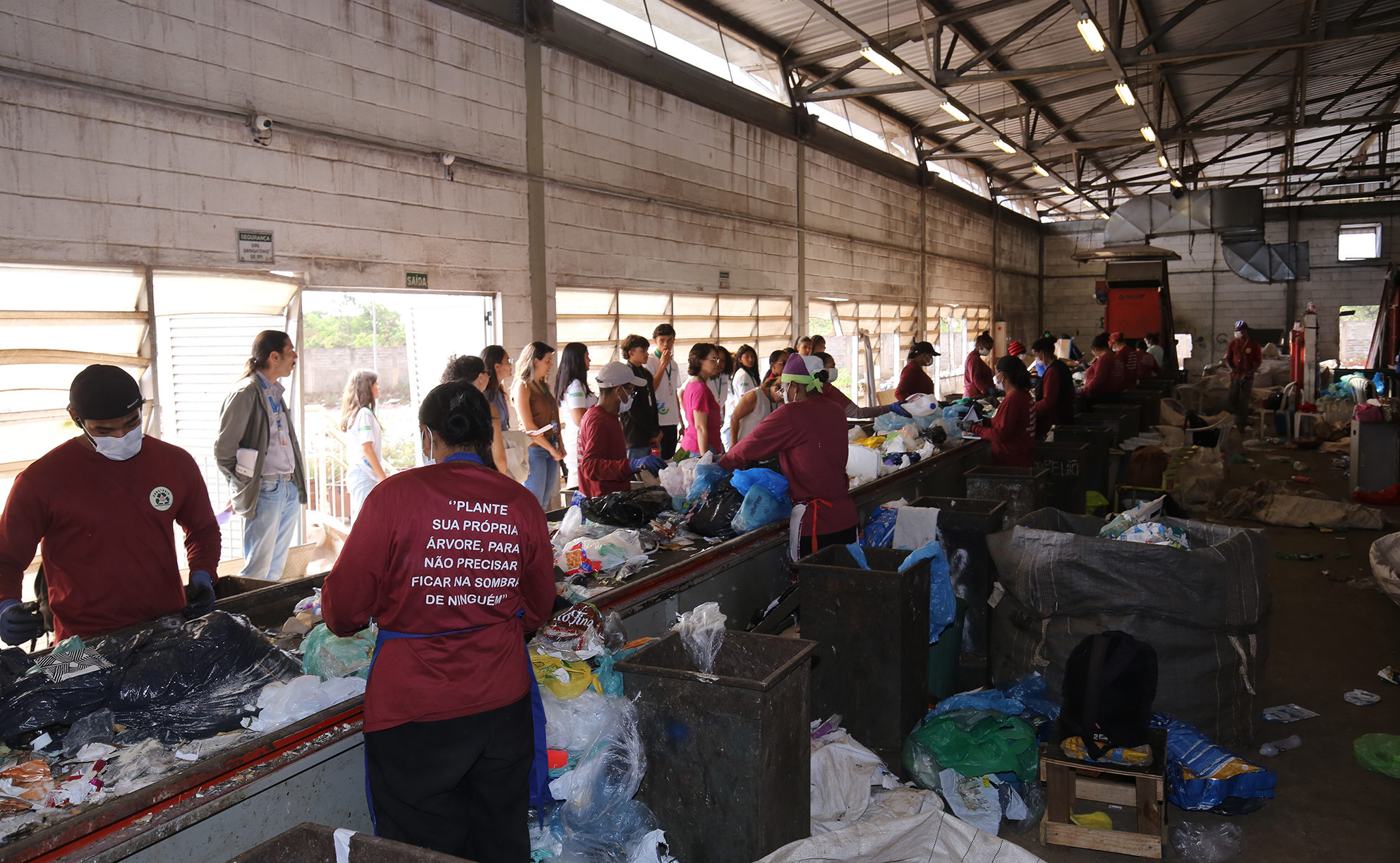 Grupo de trabalhadores com uniformes vermelhos realiza a triagem de materiais recicláveis em uma esteira dentro de um galpão industrial.