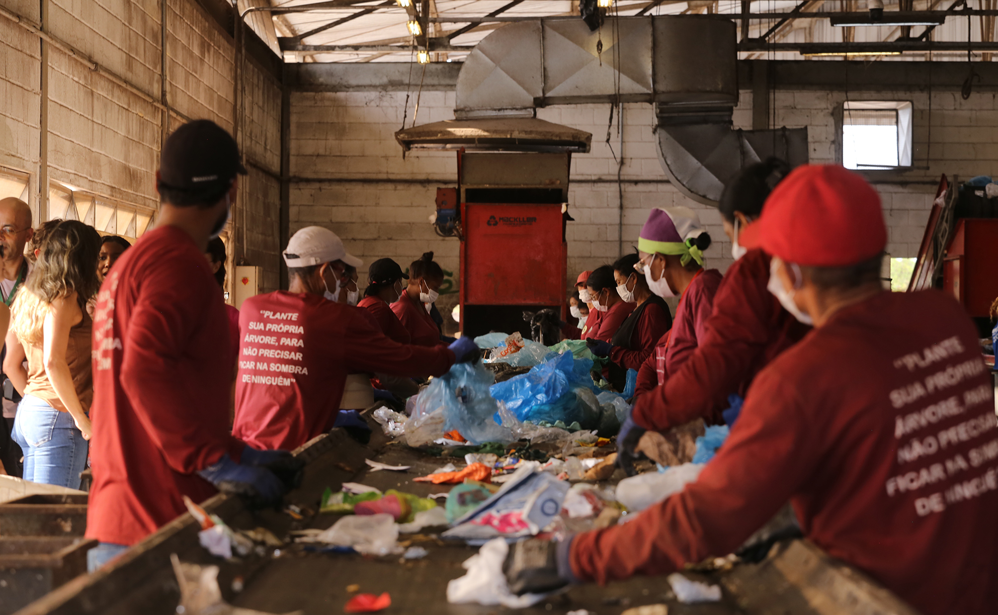 Trabalhadores com camisetas vermelhas e luvas de proteção fazem a triagem de resíduos recicláveis em uma esteira dentro de um galpão.